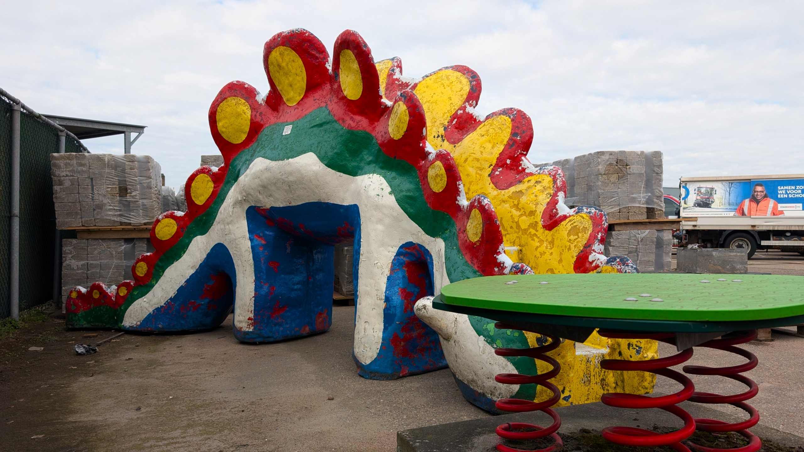 Speeltoestel in de vorm van een kleurrijke dinosaurus met rode, gele en groene tinten op een industrieterrein.