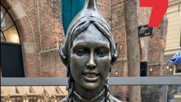 Een bronzen beeld van een vrouw met een puntige hoed en vlechtjes, geplaatst tegen een achtergrond van bakstenen gebouwen.