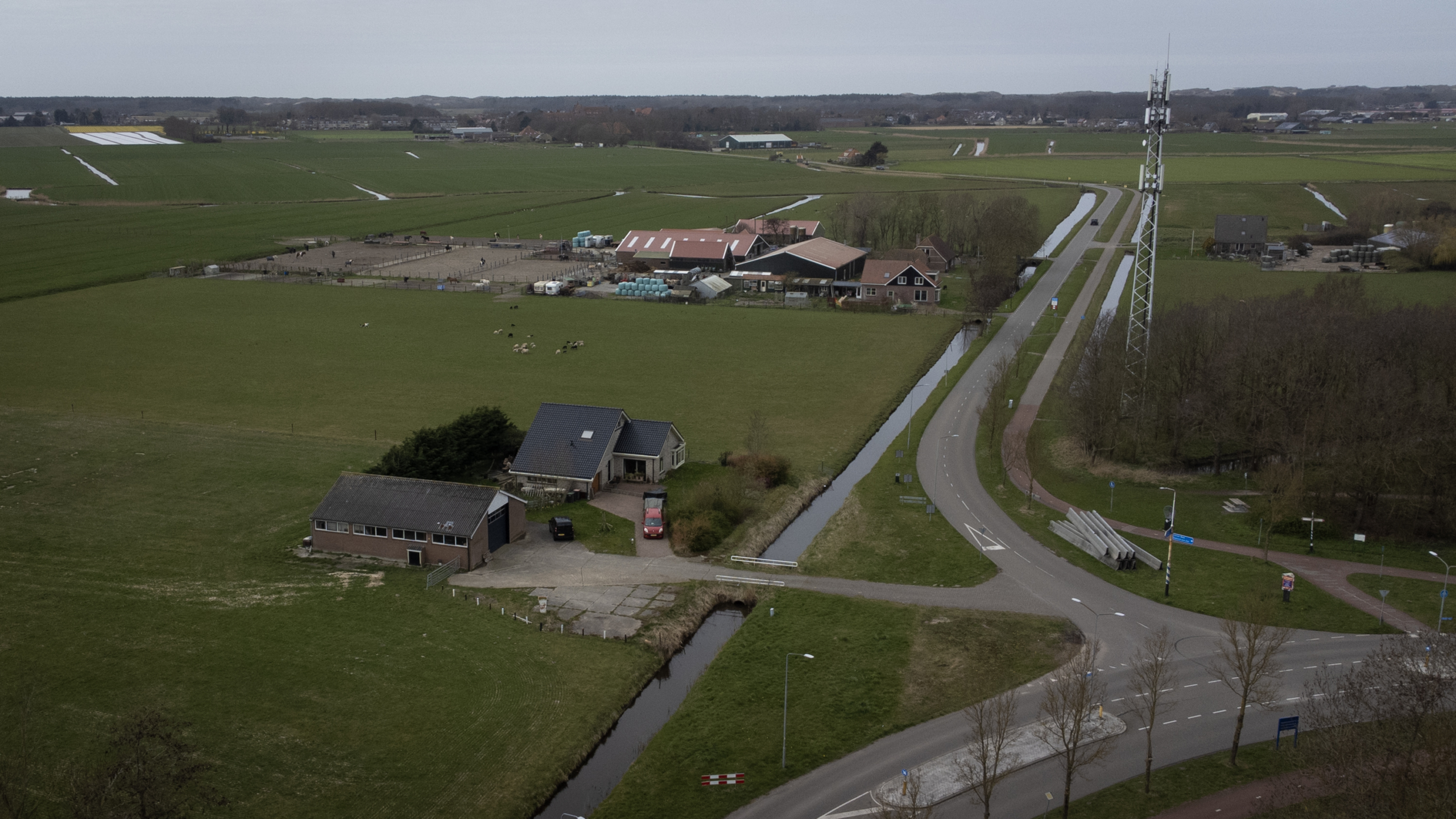 Landelijk gebied met boerderij, weilanden, een weg en een zendmast, omgeven door kanalen en verspreide bomen.