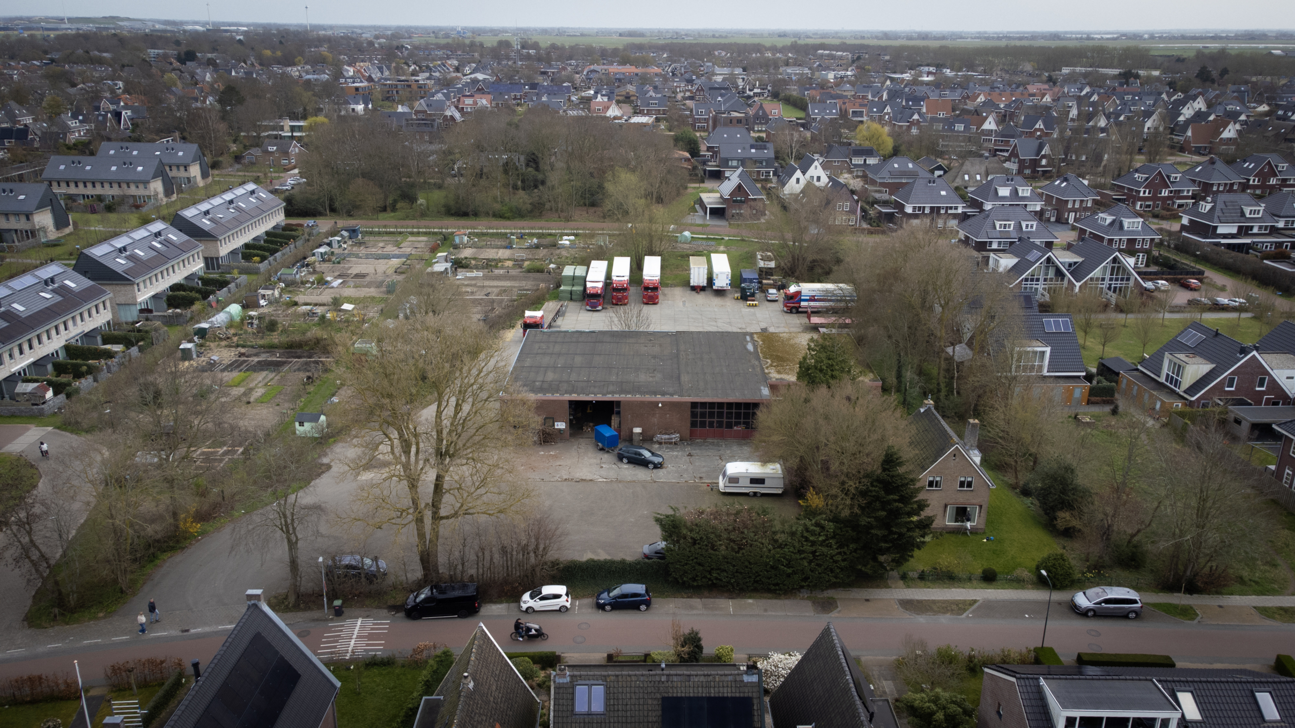 Luchtfoto van een woonwijk met huizen en een bedrijventerrein, met geparkeerde vrachtwagens en auto's op de voorgrond.