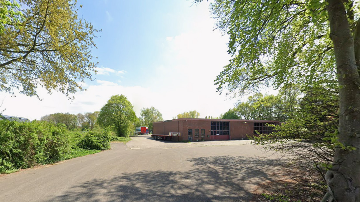 Bakstenen gebouw met grote garagedeuren, omringd door bomen en groene struiken op een zonnige dag.