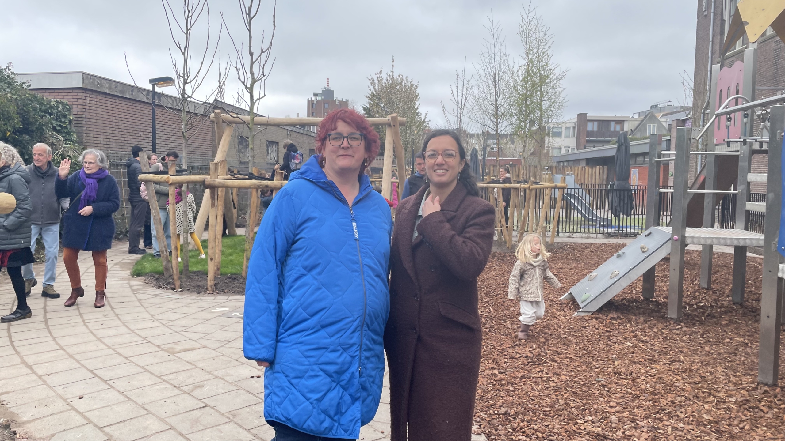 Twee vrouwen staan op een speelplaats, omringd door andere mensen en spelende kinderen.