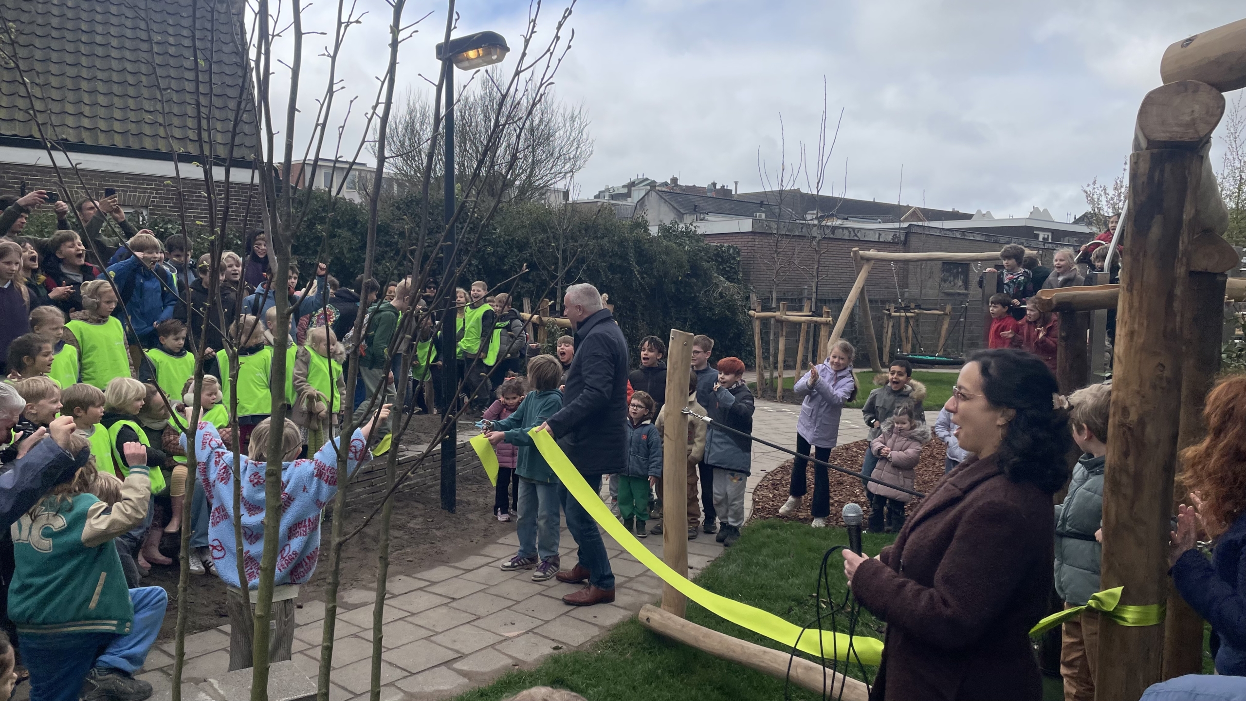 Een groep kinderen en volwassenen viert enthousiast de opening van een speeltuin terwijl een lint wordt doorgeknipt.