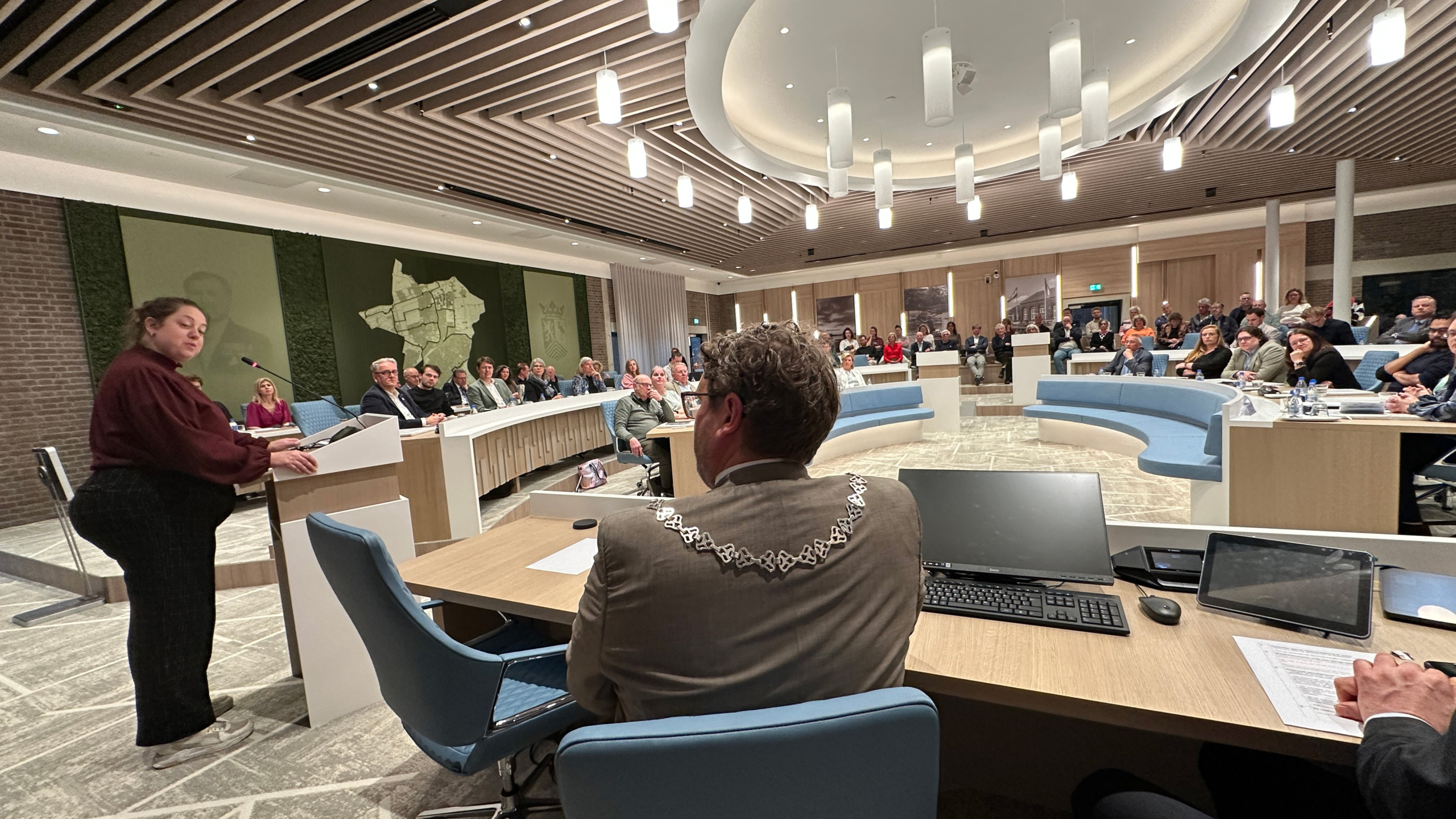 Een vrouw spreekt in een moderne vergaderzaal vol met mensen, met een persoon op de voorgrond die een ambtsketen draagt.