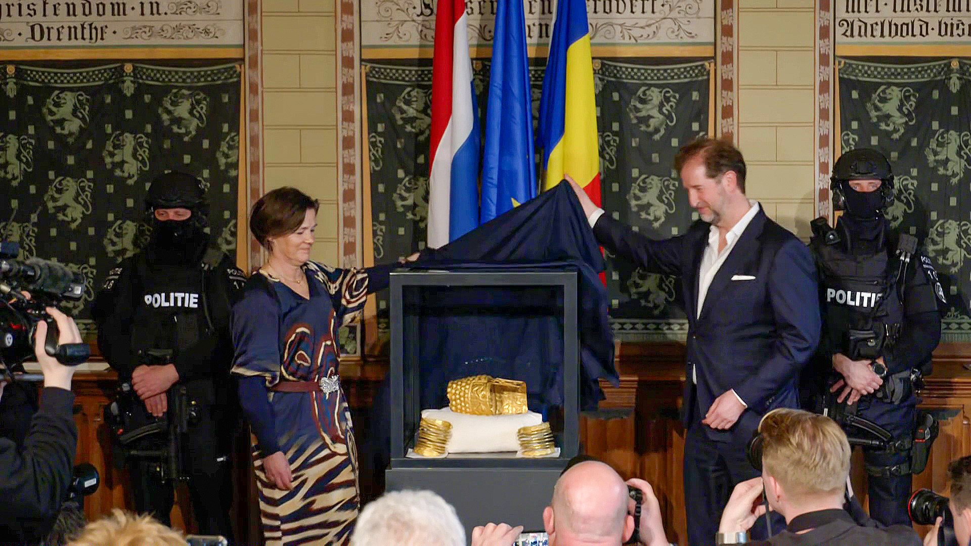Een man en een vrouw onthullen een vitrine met gouden artefacten, omringd door beveiligingsagenten en persfotografen.