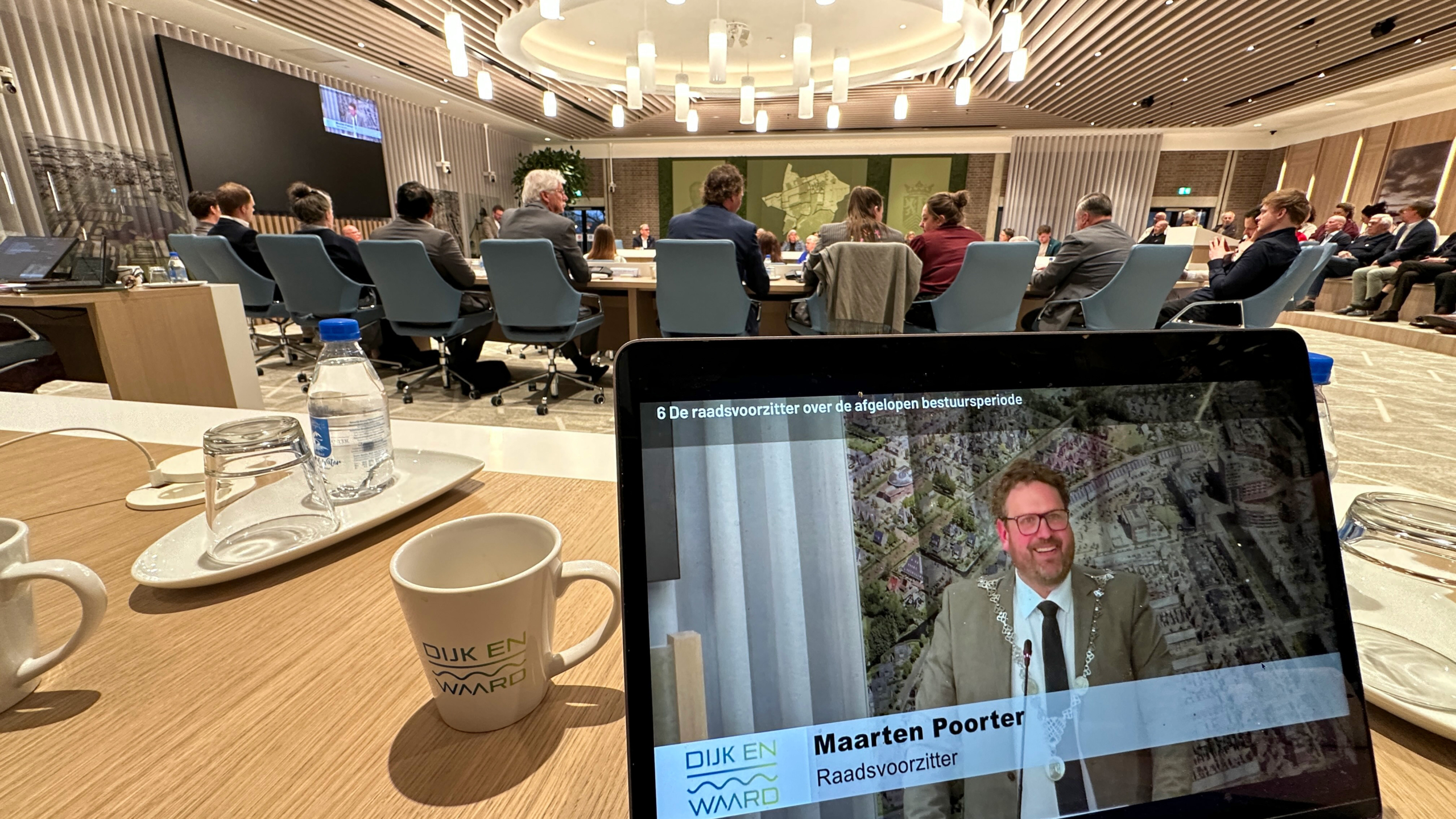 Vergaderruimte met mensen aan een tafel; op een laptop is een presentatie te zien met de tekst "Maarten Poorter Raadsvoorzitter".