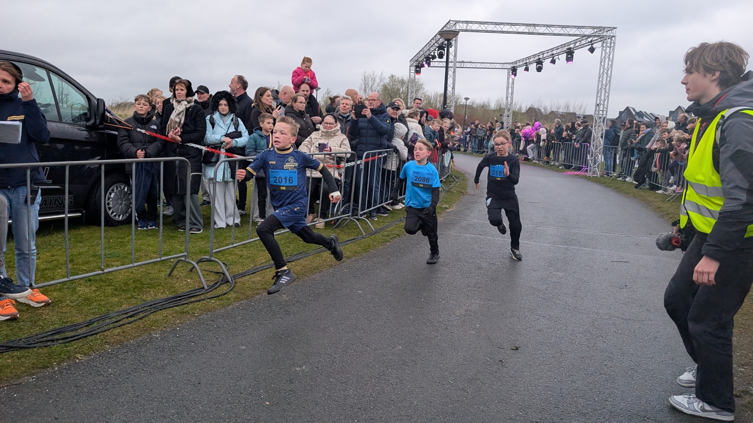 Drie kinderen rennen een hardloopwedstrijd, terwijl toeschouwers achter hekken langs de weg staan te kijken.