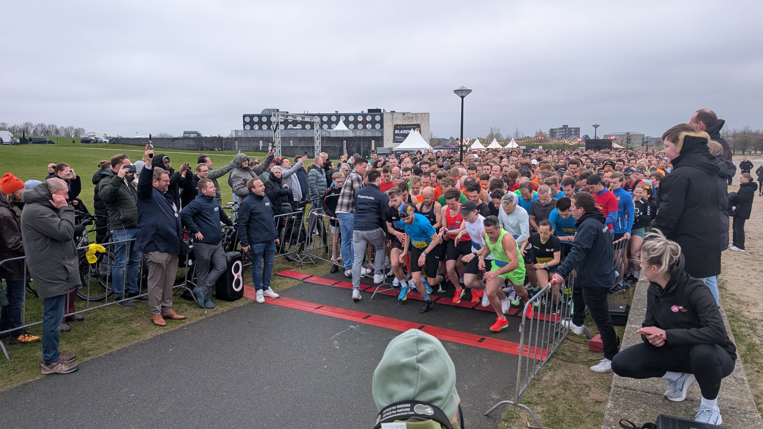 Een menigte hardlopers staat klaar aan de startlijn van een race, omringd door toeschouwers en organisatoren.