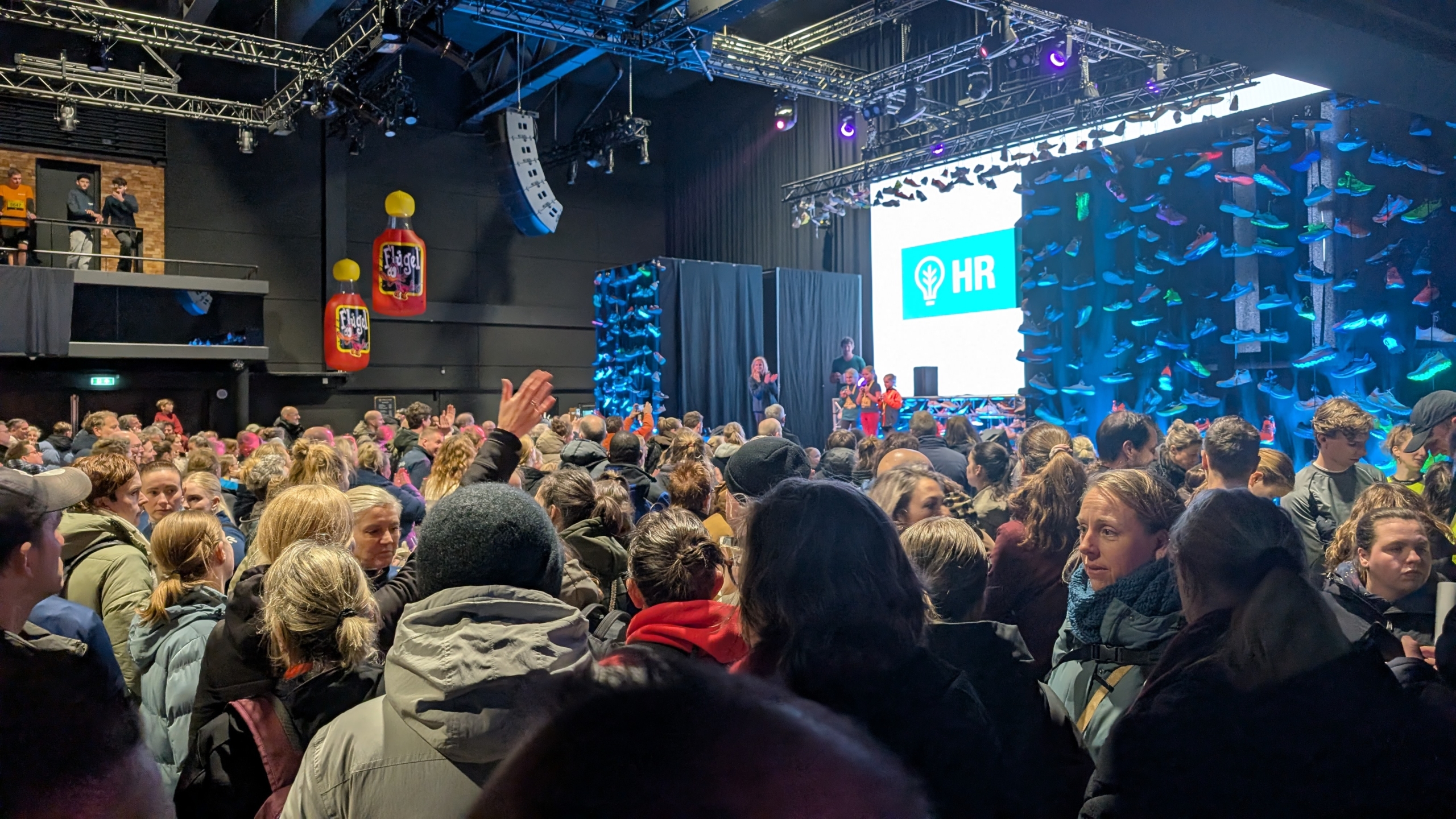 Een grote menigte verzamelt zich in een zaal met opgehangen schoenen, terwijl op het podium een scherm met "HR" zichtbaar is.