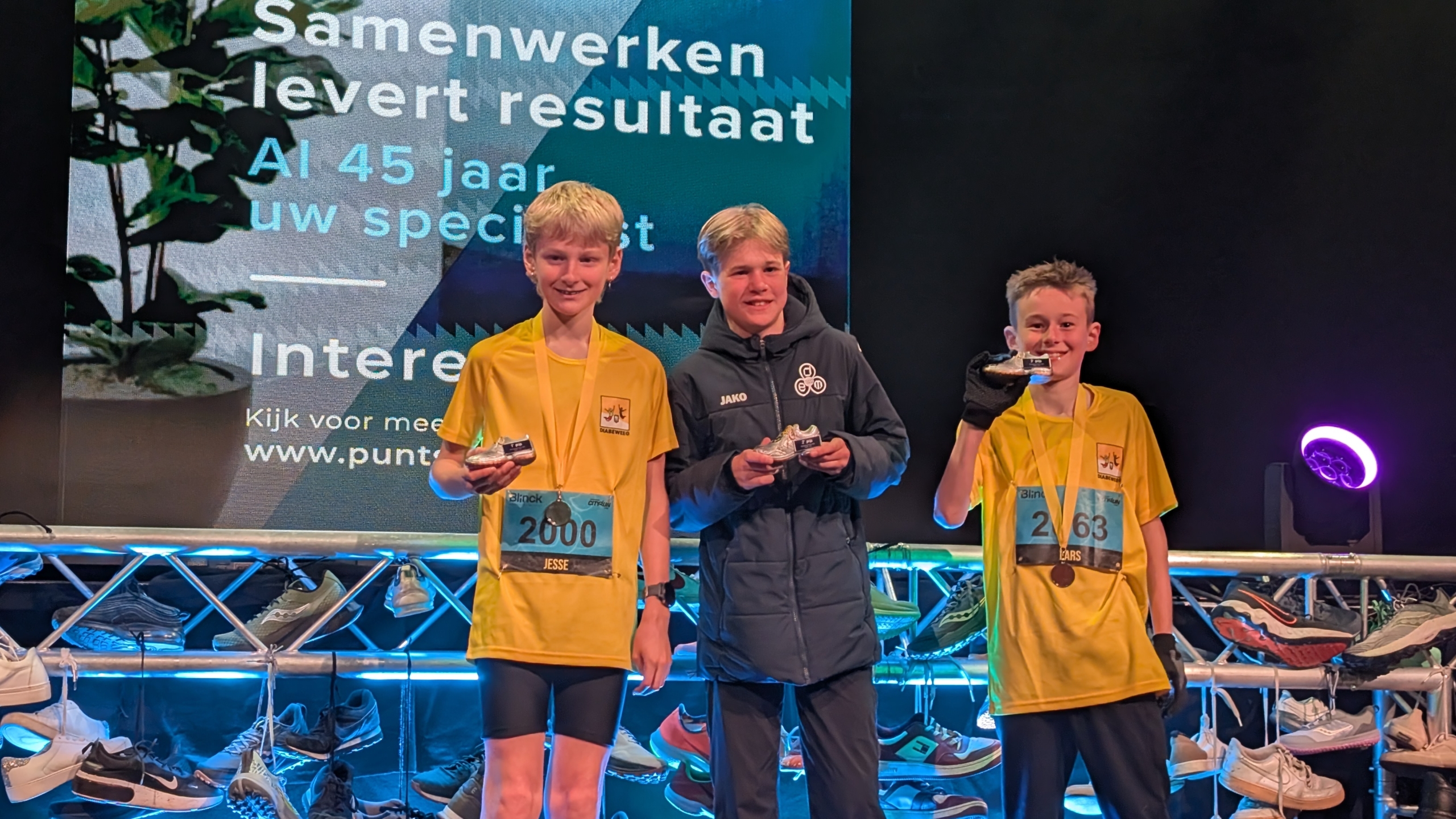 Drie jonge jongens poseren met medailles voor een bord met tekst, tijdens een hardloopevenement.