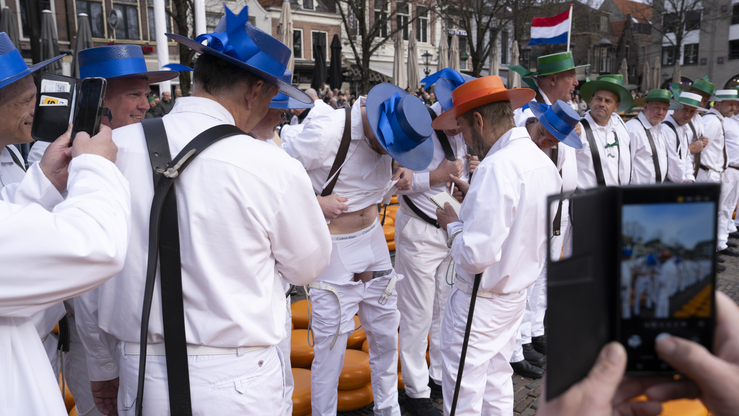 Mannen in witte kleding met gekleurde hoeden, bezig met een ceremonie tussen gestapelde kazen in een Nederlandse stad.
