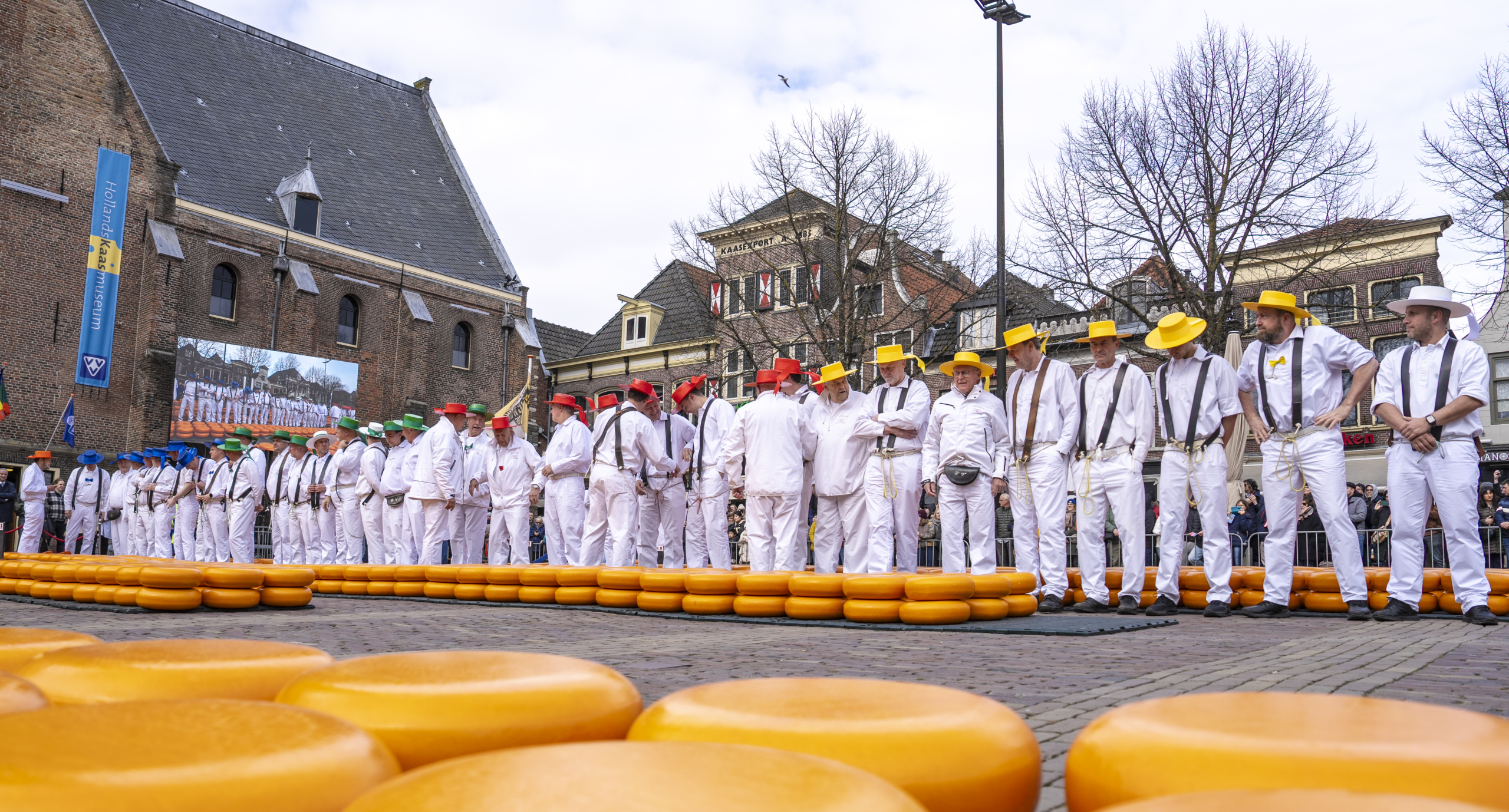 Mannen in witte kleding en gekleurde hoeden staan bij stapels kaas op een plein bij een kaasmarkt.