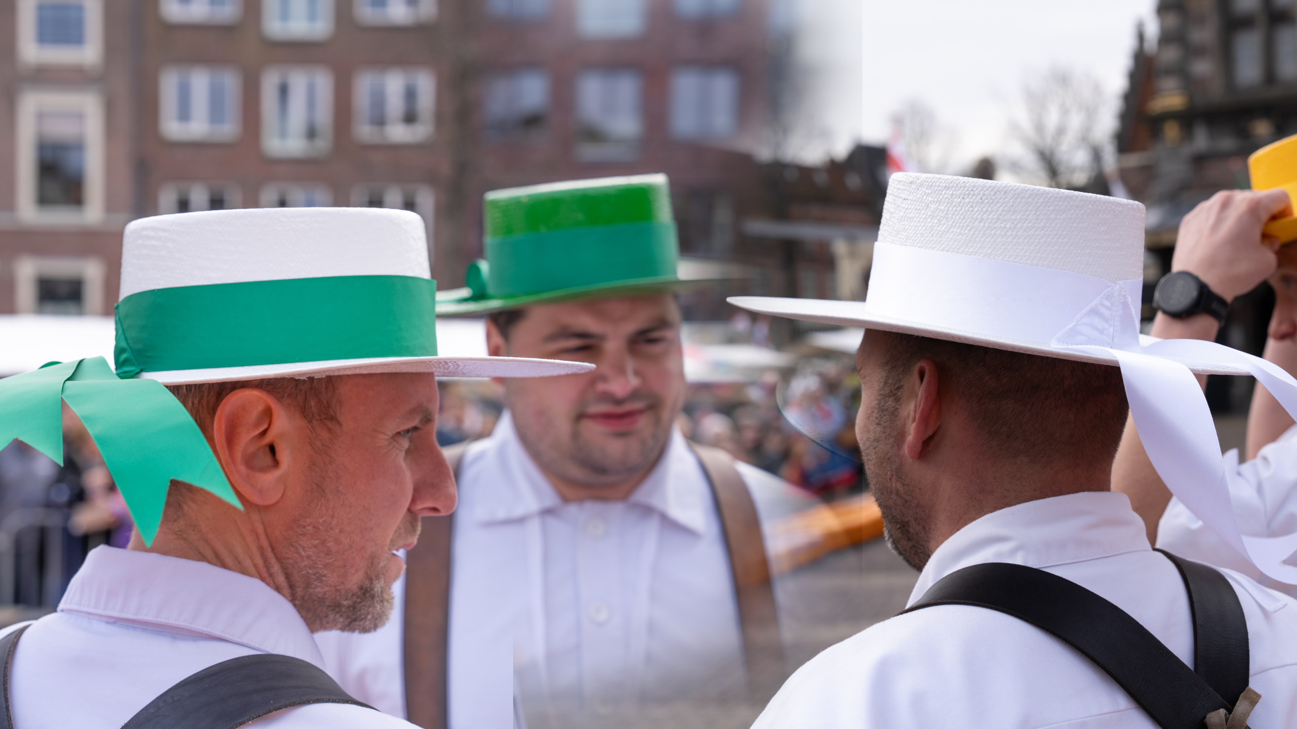 Mannen in witte kleding en hoeden met groene linten praten buiten, met een vage stad op de achtergrond.