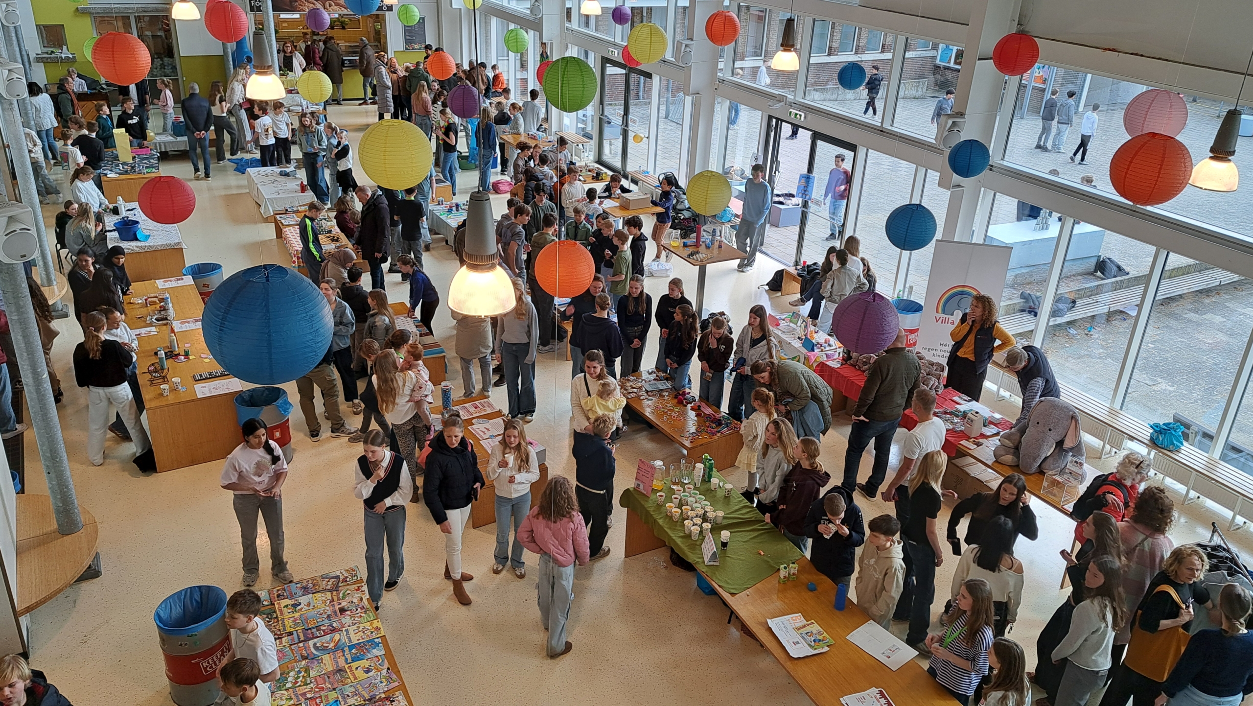 Een drukbezochte schoolbeurs met kleurrijke lampionnen, kraampjes en bezoekers, waaronder veel kinderen en volwassenen.