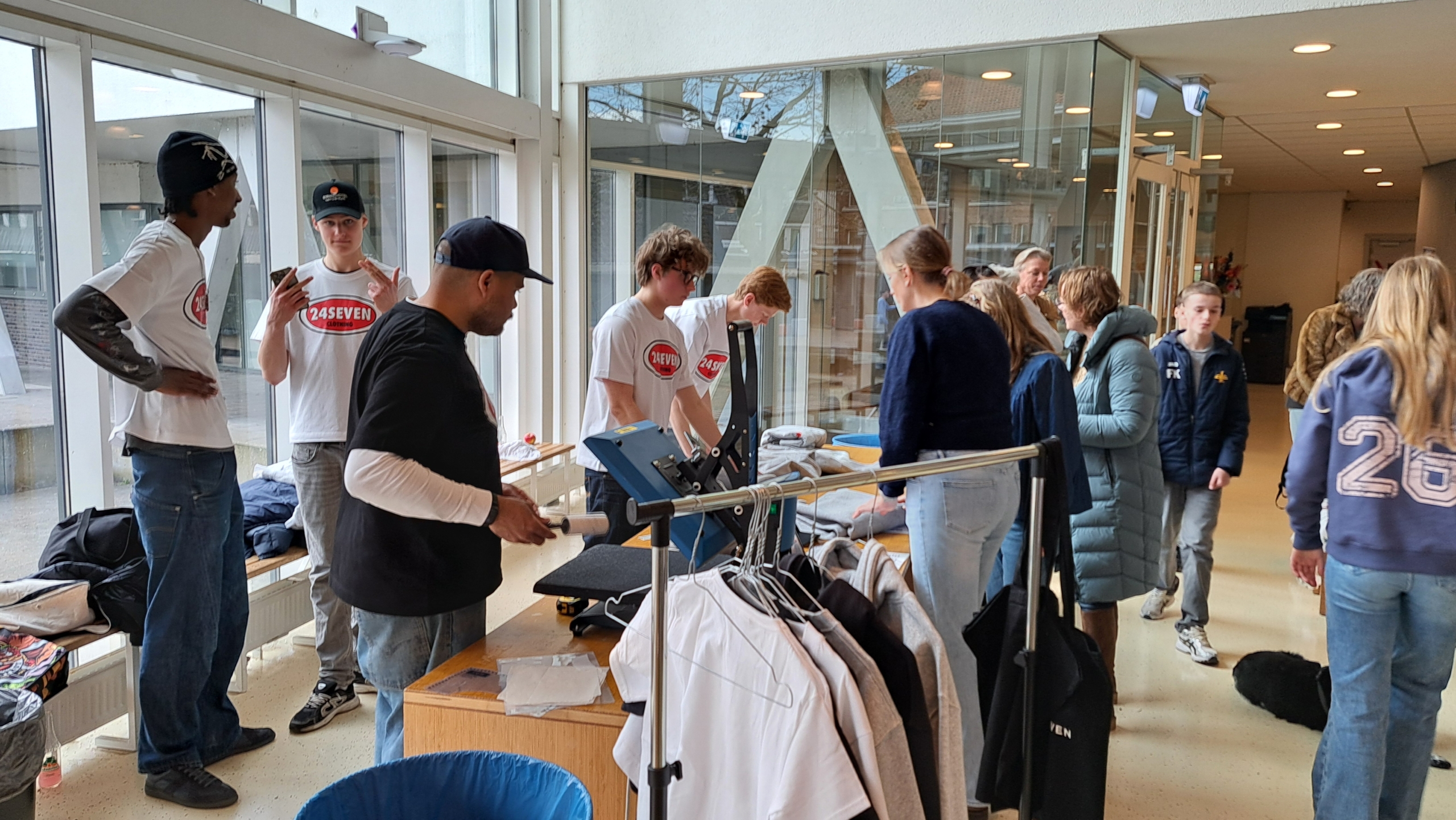 Mensen in een lichte ruimte met glazen wanden, bezig met kleding, sommige in shirts met 24SEVEN-logo.
