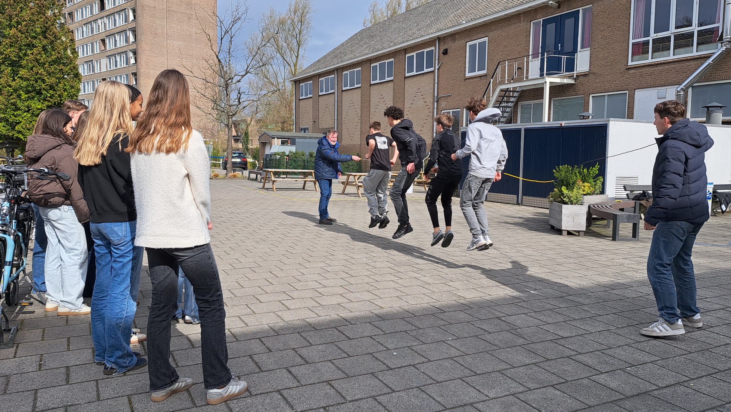 Een groep jongeren en een man springen samen in een spel op een plein met fietsen en een gebouw op de achtergrond.
