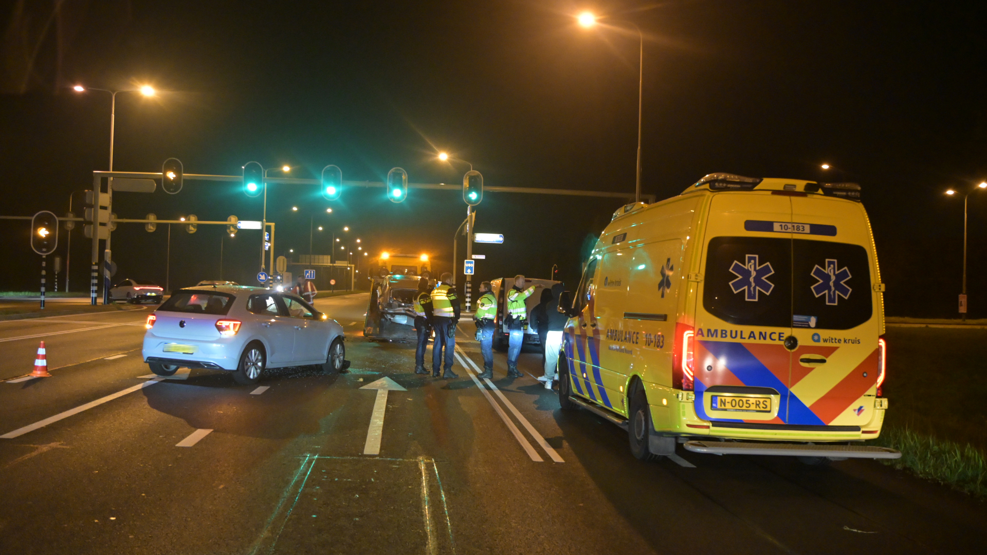 Verkeersongeluk in de nacht bij een kruispunt; ambulance en verschillende auto's aanwezig met politie en verkeerslichten op groen.