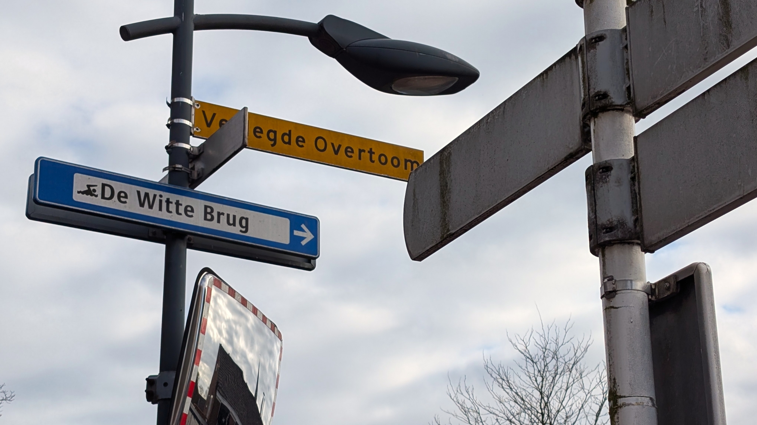 Straatnaamborden met "Voetwegde Overtoom" en "De Witte Brug" bij een lantaarnpaal onder een grijze lucht.