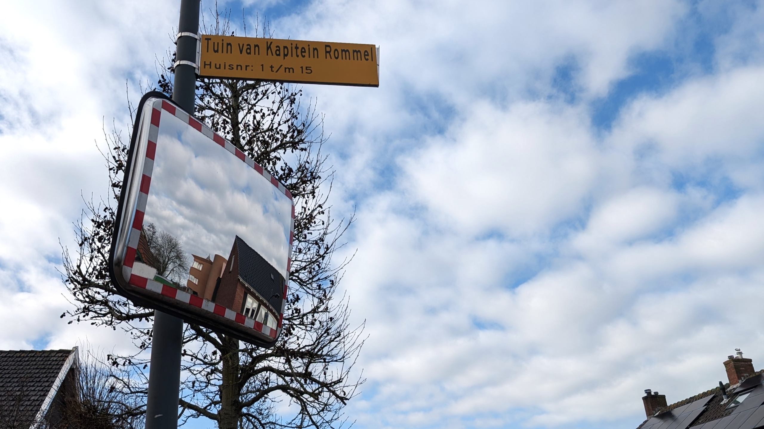 Verkeersspiegel met weerspiegeling van huizen onder een bordje "Tuin van Kapitein Rommel" tegen een bewolkte hemel.