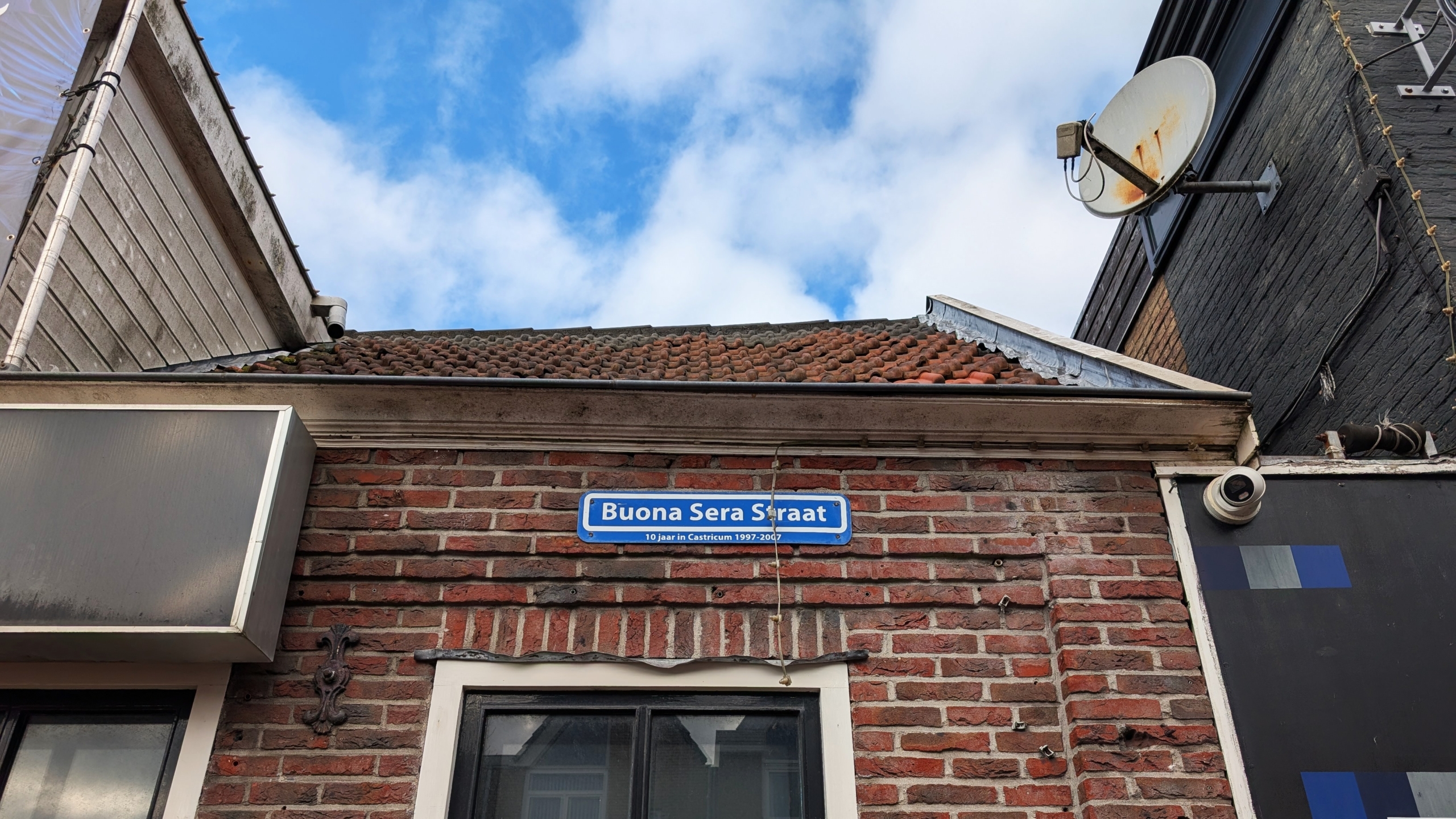 Straatnaambord met de tekst "Buona Sera Straat" op een bakstenen gevel, met een schotelantenne rechtsboven.