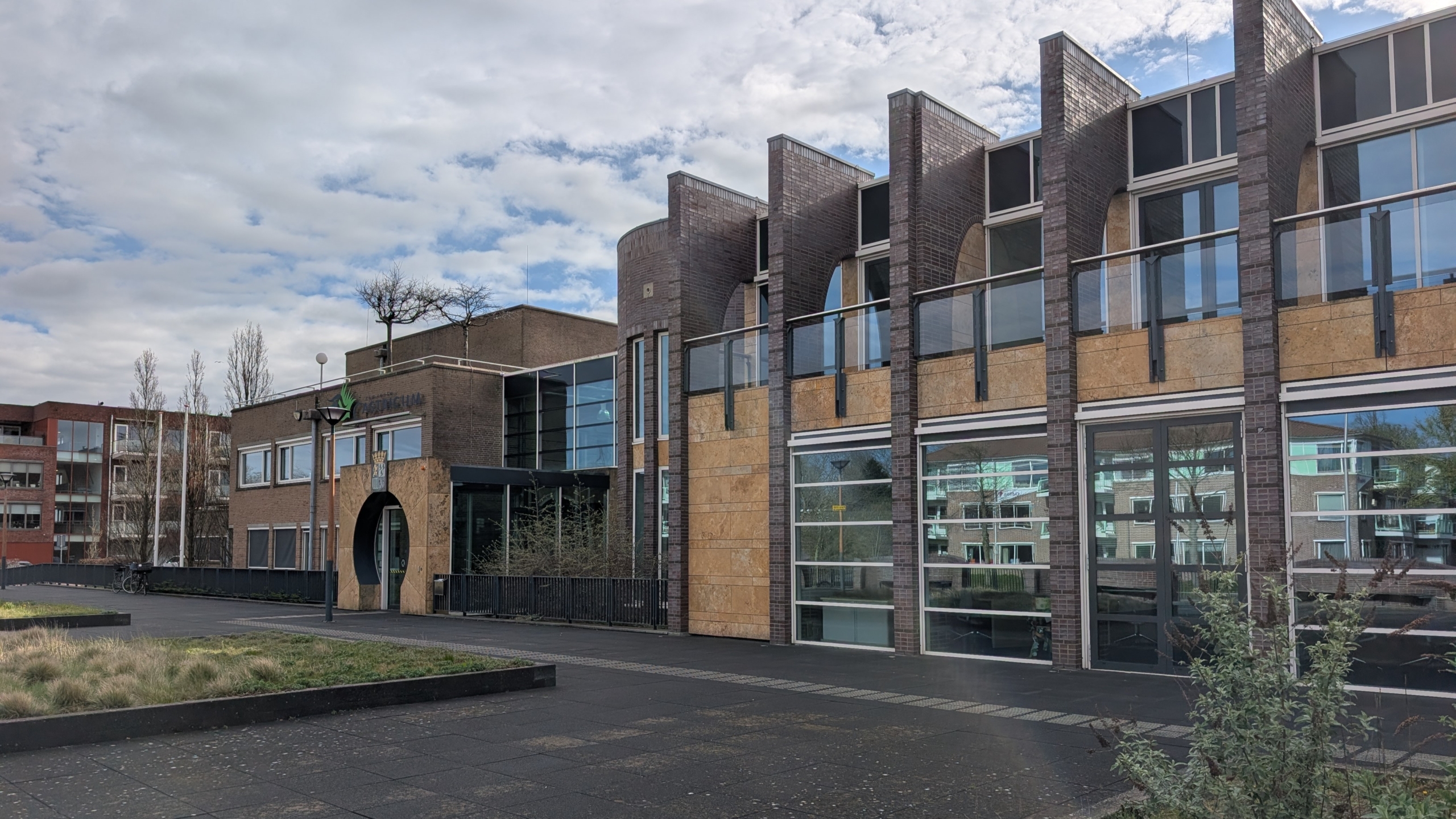 Modern gebouw met grote ramen, bakstenen facade en geometrische vormen onder een bewolkte hemel.