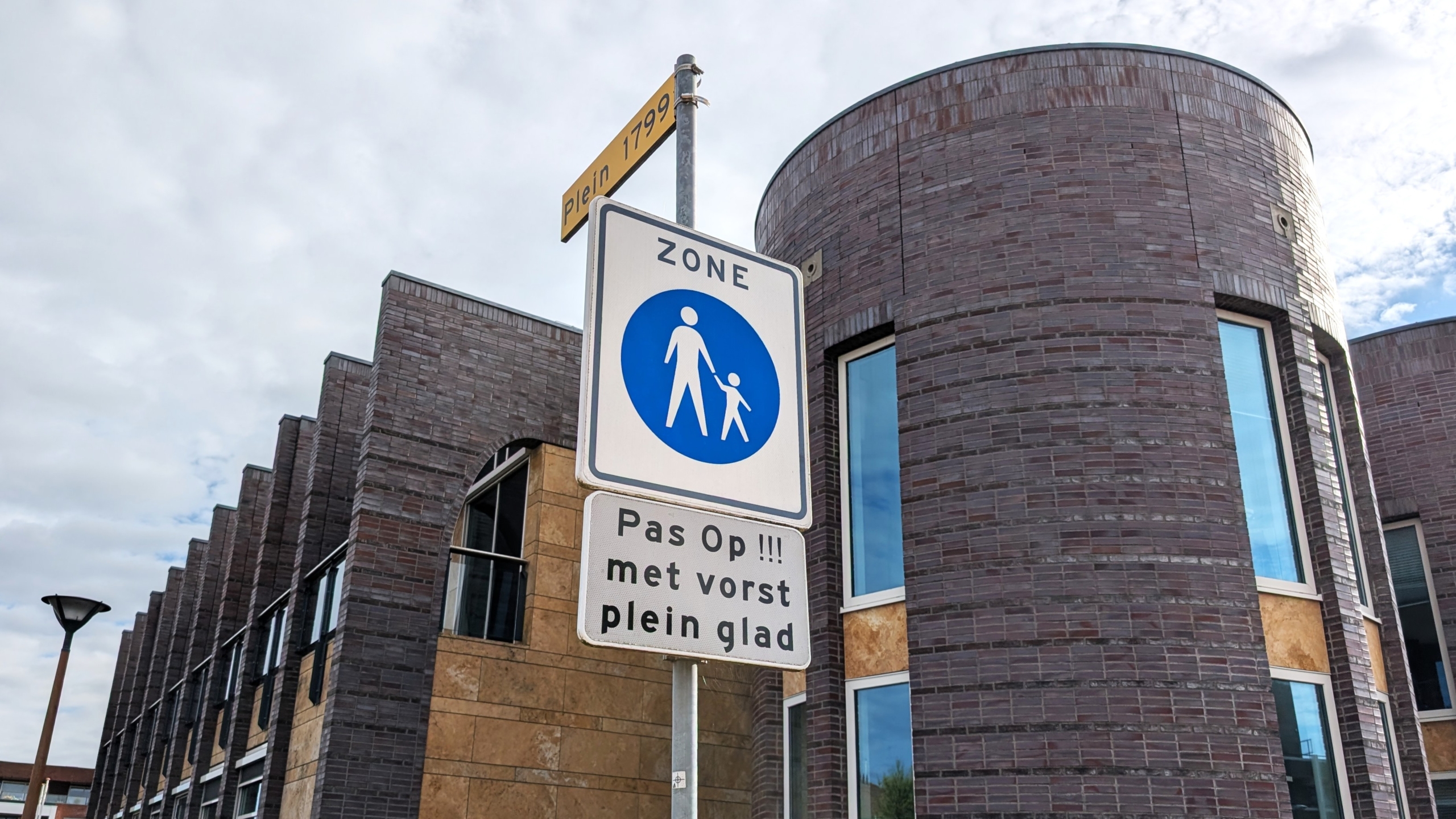 Verkeersbord met waarschuwing voor glad plein bij vorst, tegen de achtergrond van een modern, rond stenen gebouw.
