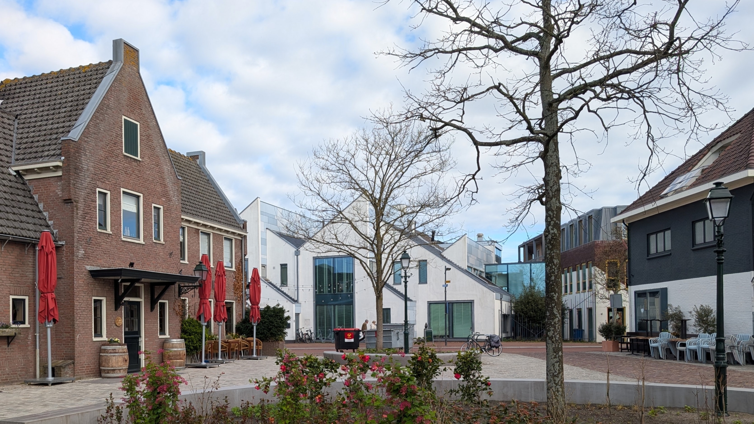 Plein met oude en moderne gebouwen, een kale boom, rode parasols, rozenstruiken en een vuilnisbak.