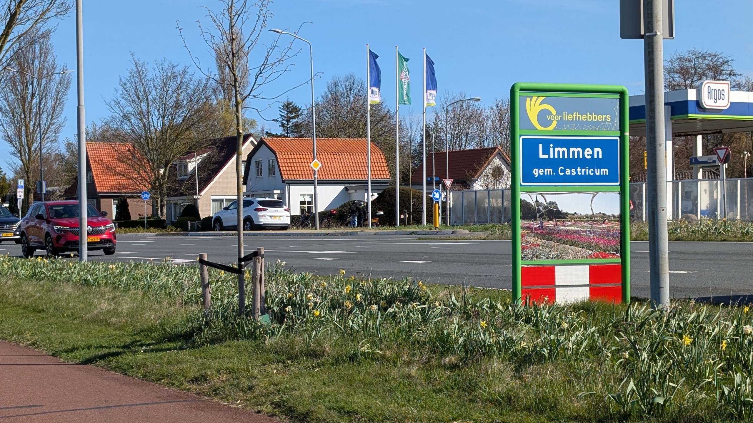 Bord bij een rotonde met "Limmen gem. Castricum" en afbeeldingen van bloemen. Op achtergrond huizen en een tankstation.