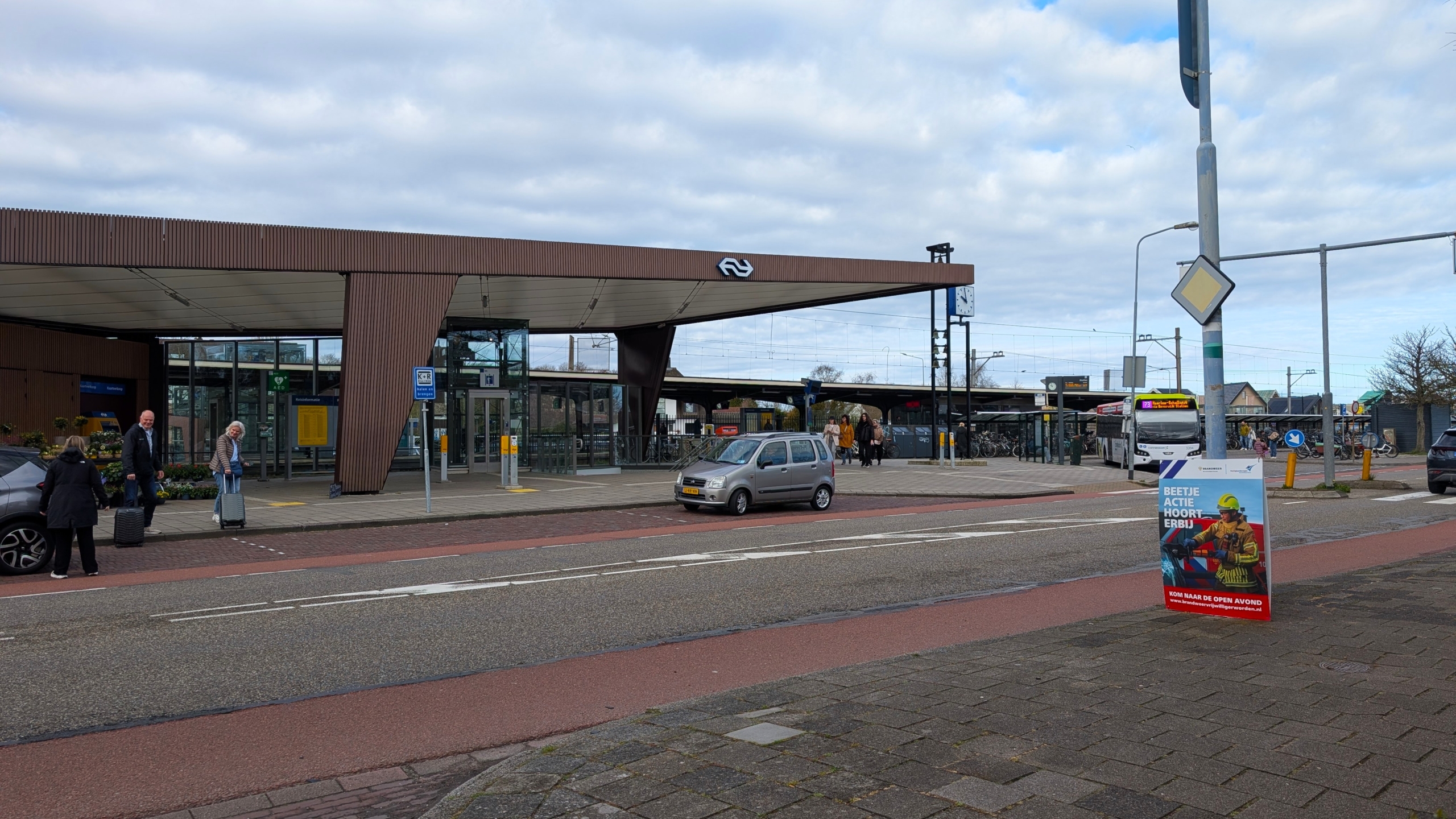 Busstation voor een treinstation met verschillende mensen en voertuigen; een reclamebord staat aan de zijkant van de straat.