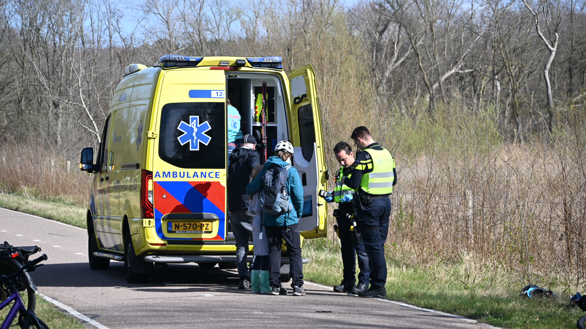 Ambulance met geopende deuren naast een weg, omringd door politieagenten en personen met fietsoutfits.