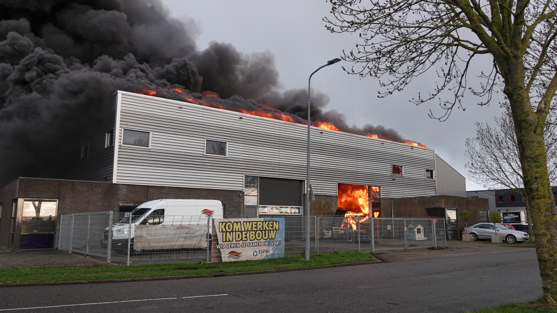 Een fabrieksgebouw in brand met dikke zwarte rook en vlammen die uit het dak en de ramen komen.