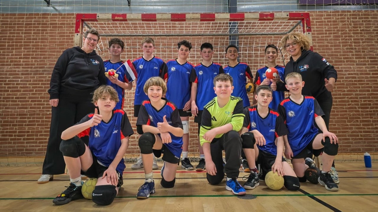 Jeugdhandbalteam poseert voor een doel met coaches, in blauwe shirts, enkele kinderen houden handballen vast.