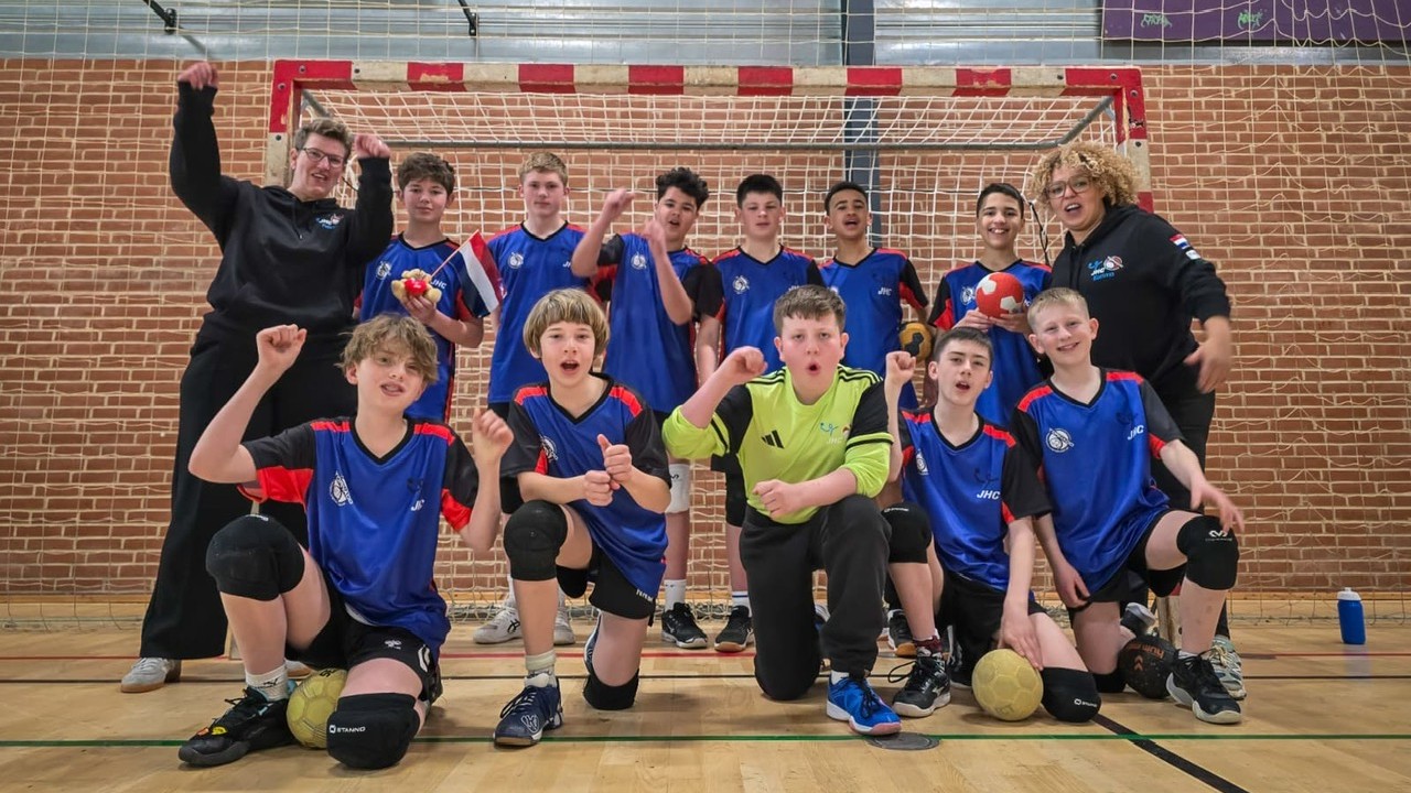 Een jeugdhandbalteam poseert enthousiast voor een doel met twee coaches, gekleed in blauwe sporttenues.