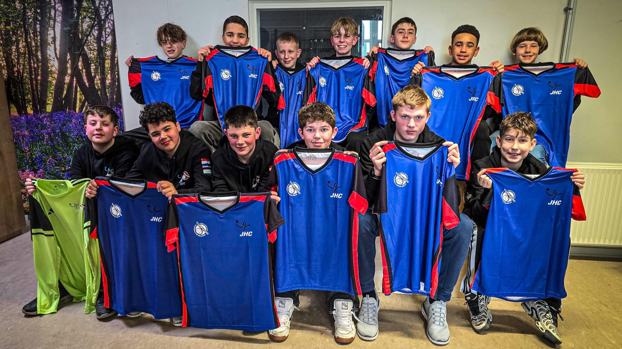 Een groep jongens poseert met blauwe en rode sportshirts met het logo "JHC" in een binnenruimte.