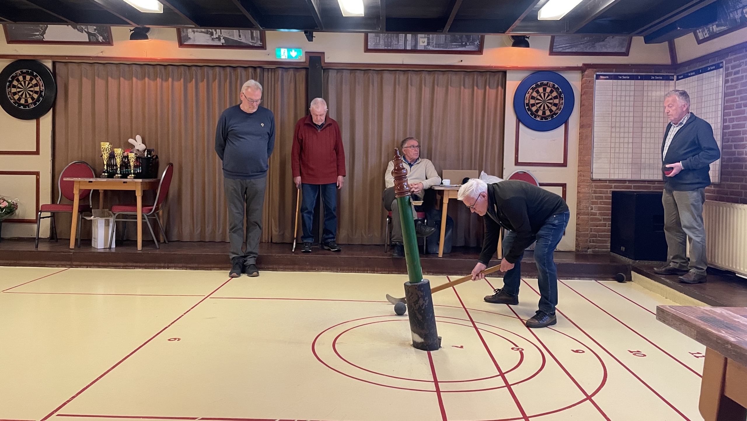 Mensen spelen doorlopend sjoelen in een zaal met dartborden en trofeeën.