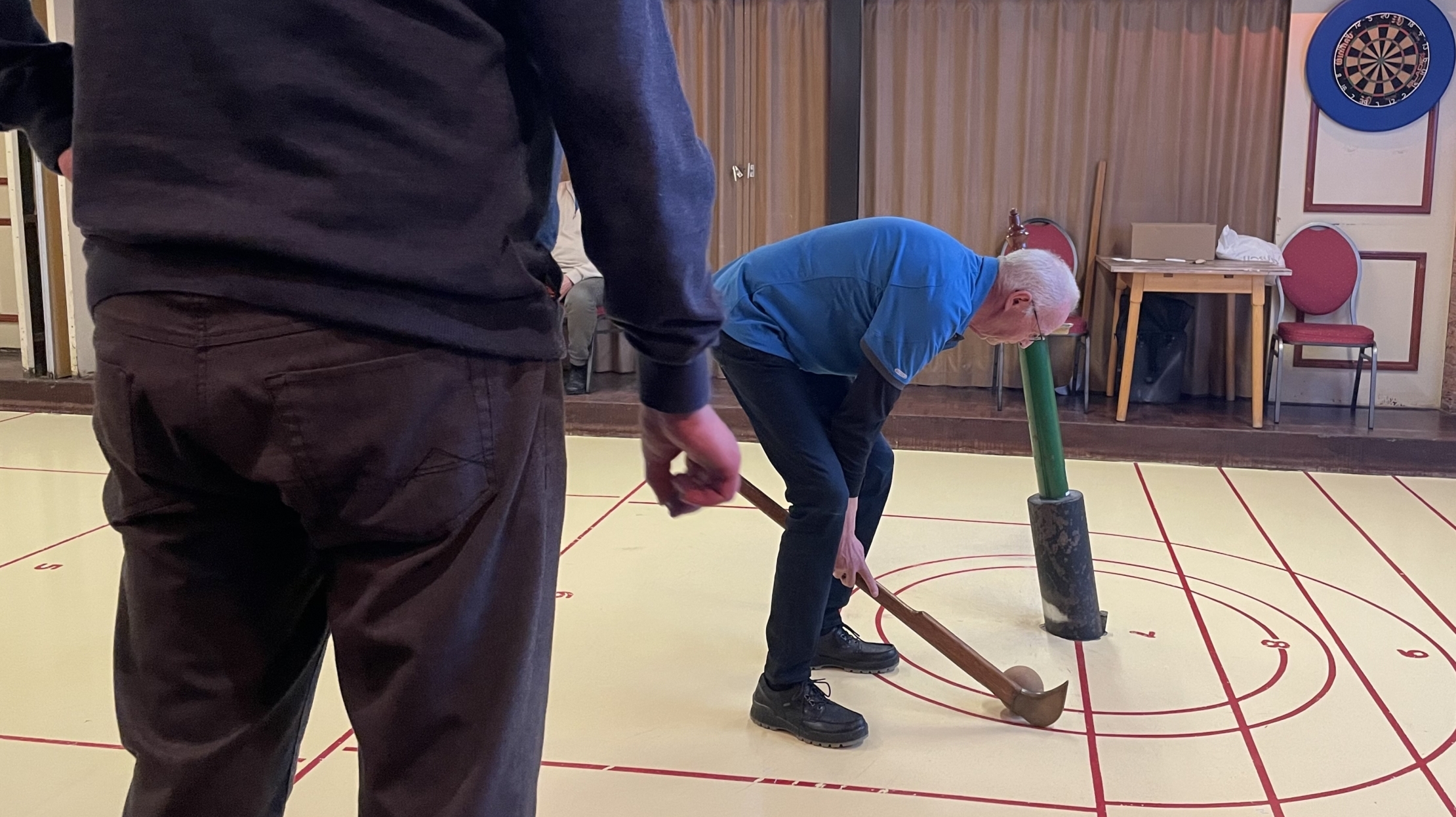 Man speelt indoor met stokken op een vloer met rode lijnen, naast een dartbord en scorebord.