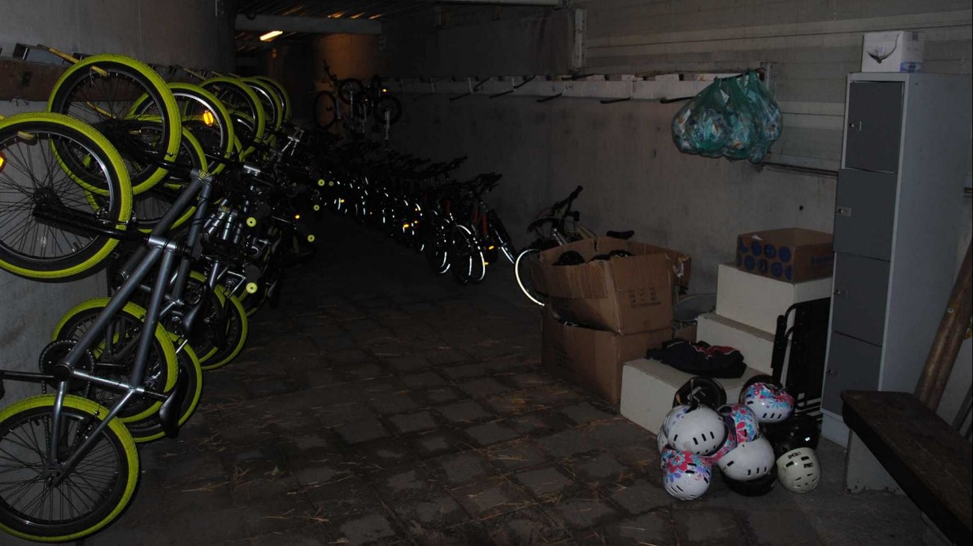 Opslagruimte met fietsen met felgroene banden, dozen, helmen, en een kast aan de rechterkant.