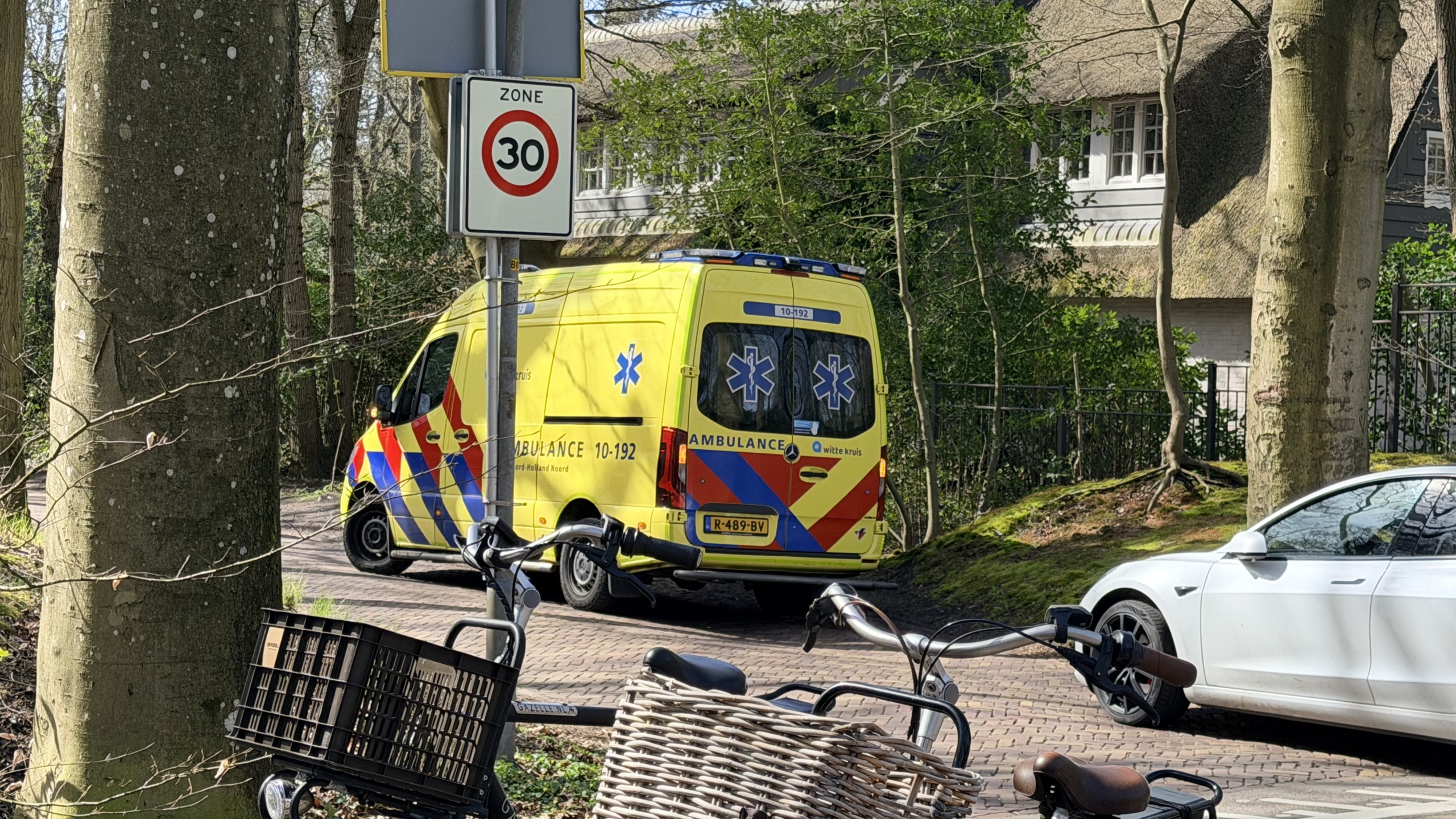 Een gele ambulance met blauwe en rode strepen staat op een weg; een verkeersbord geeft een zone van 30 km/u aan.