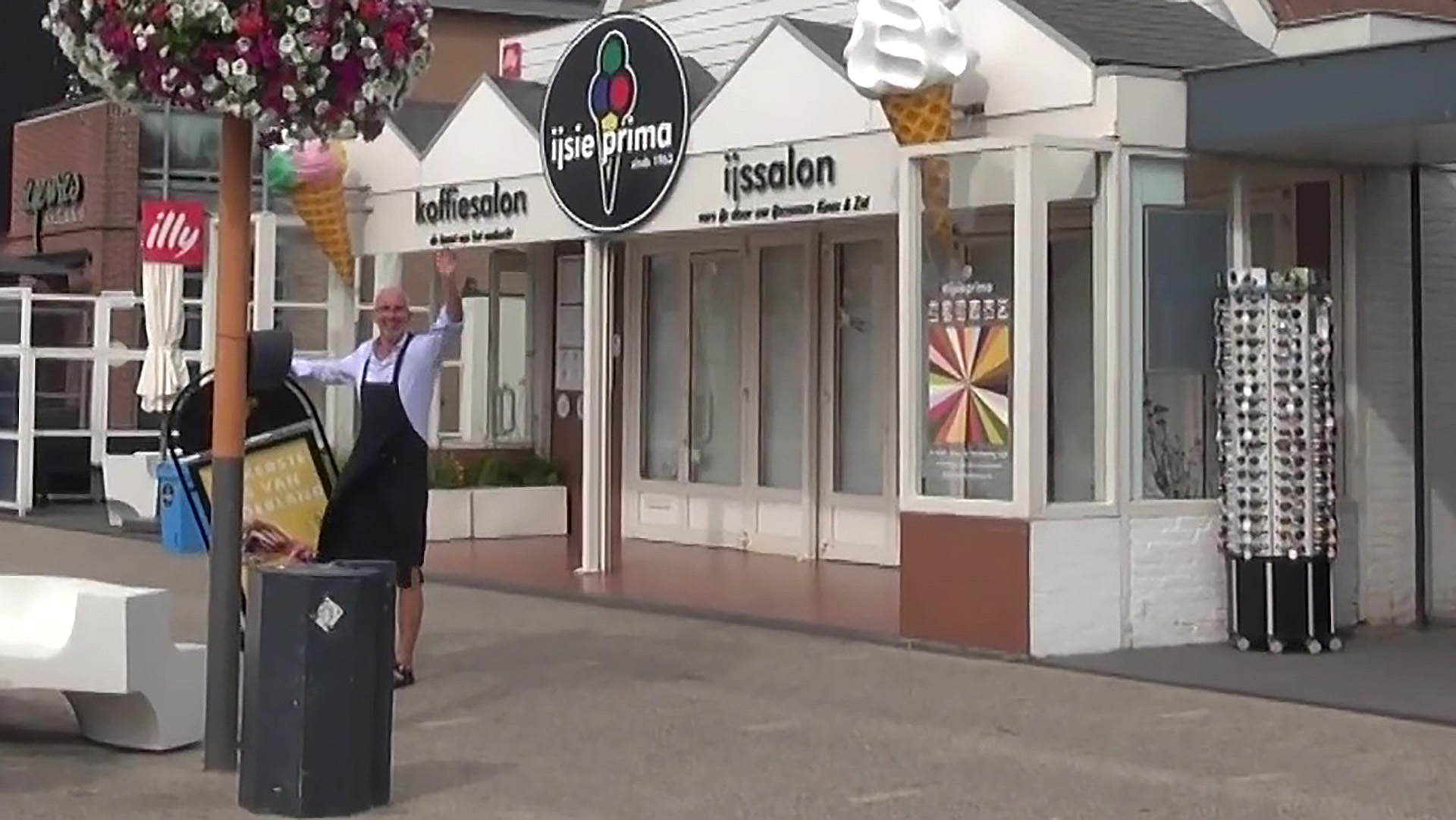 Man zwaait buiten een ijssalon met bloemenmand en opvallende ijshoorntjes als decoratie.