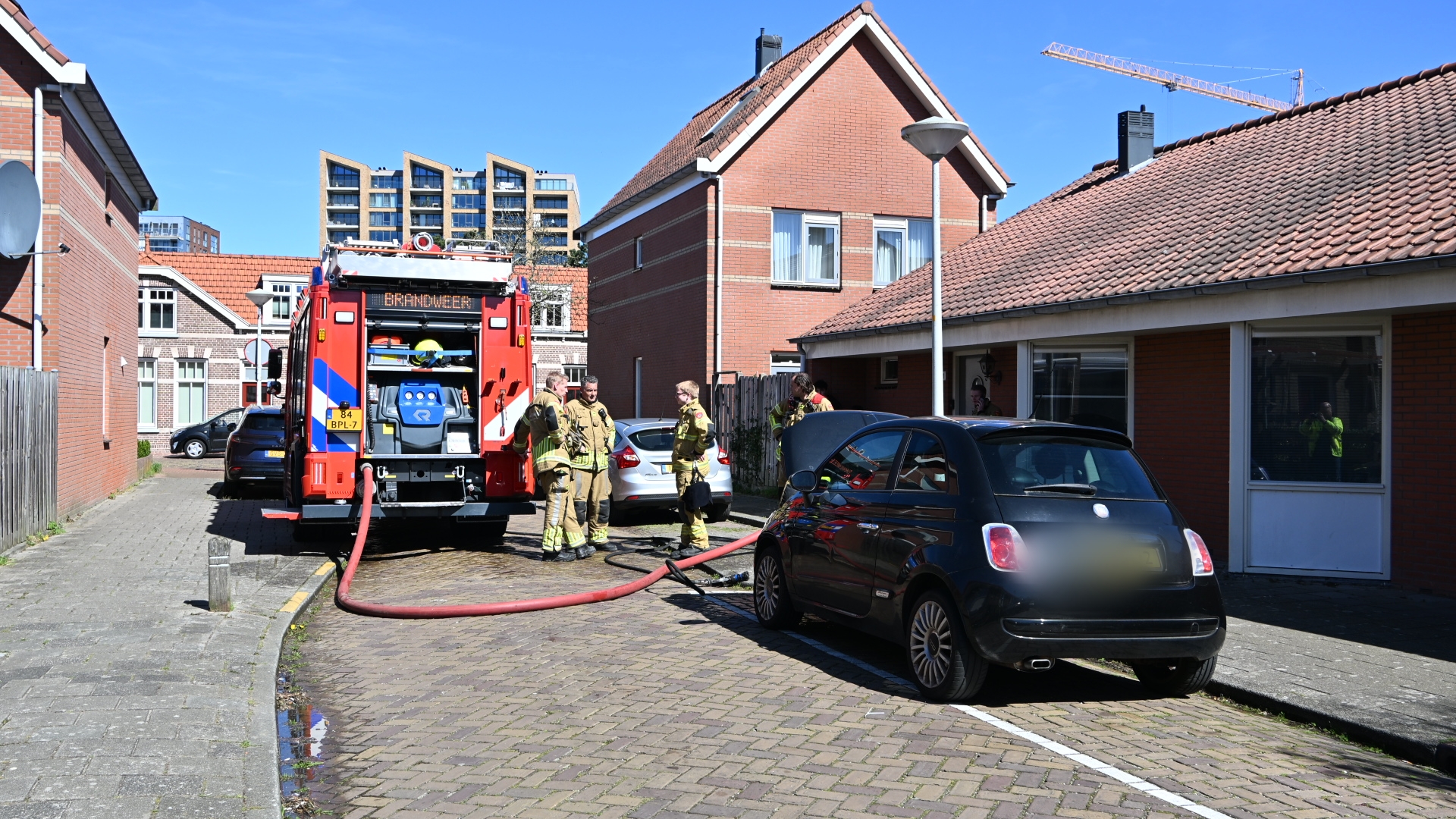 Brandweermannen naast een brandweerwagen in een woonwijk, met een brandslang aangesloten en huizen op de achtergrond.