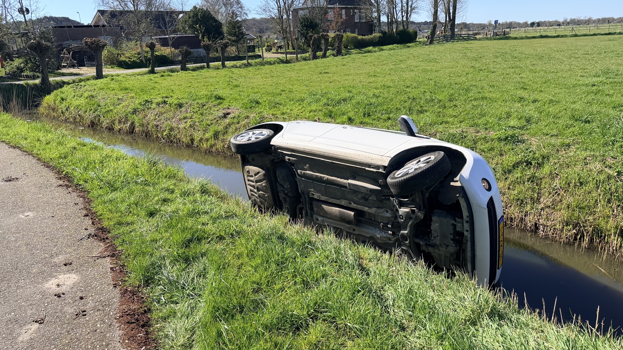 Auto op zijn kant in een sloot naast een grasveld en een weg in een landelijke omgeving.