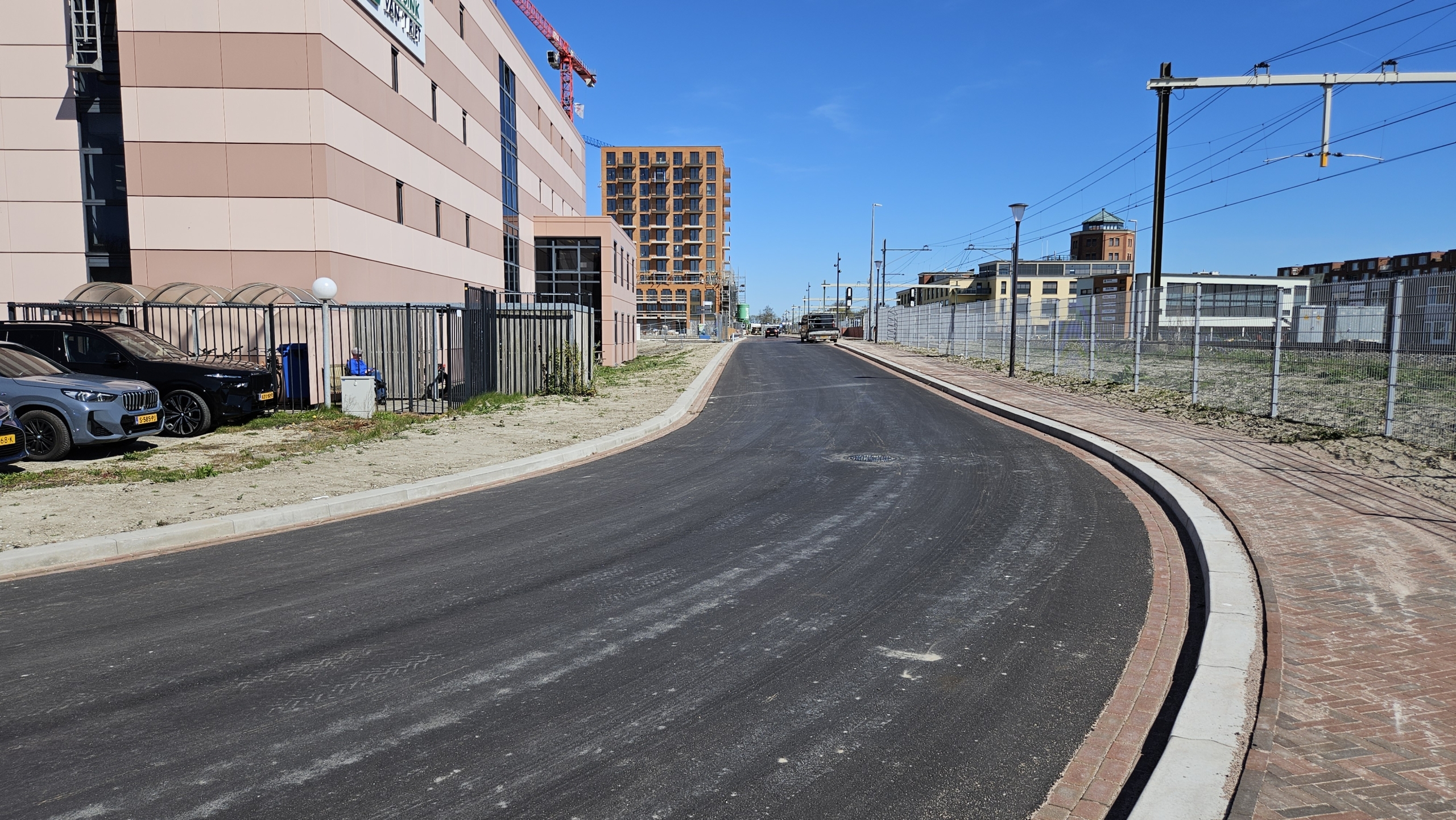 Nieuw asfalt op een weg langs een kantoorgebouw, met meerdere geparkeerde auto's en hekken aan beide zijden.