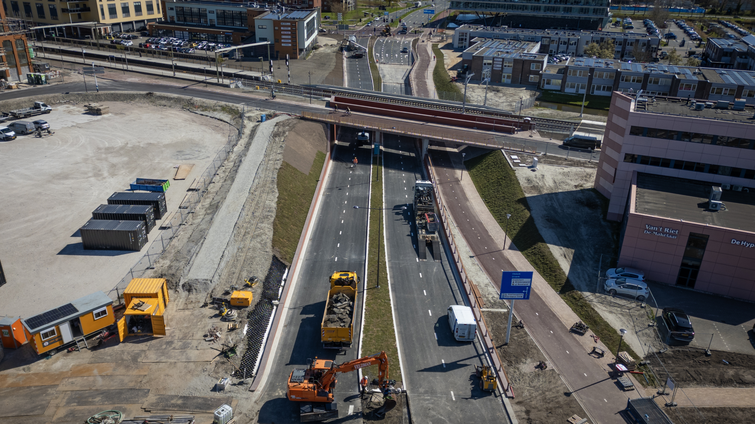 Wegwerkzaamheden aan een dubbele rijbaan met graafmachines en vrachtwagens, naast gebouwen en een brug.
