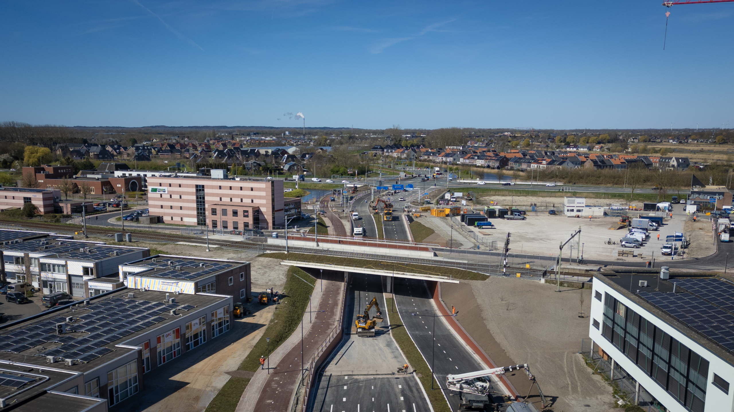 Luchtfoto van een bouwterrein met wegen en gebouwen, omgeven door een dorp en open landschap onder een heldere hemel.