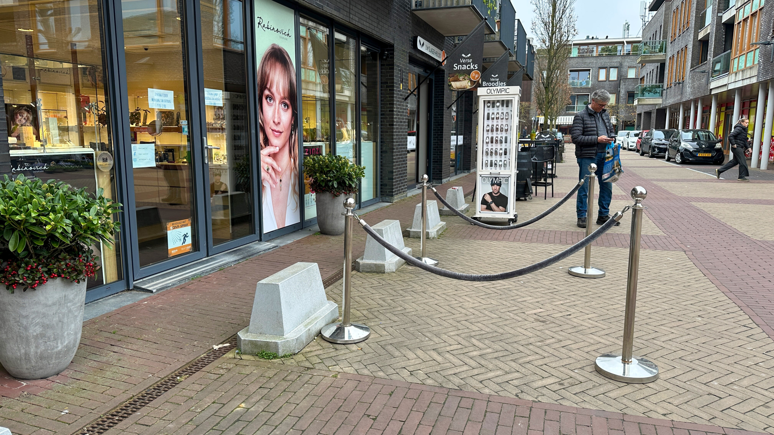 Winkelstraat met een etalage, een groot portret van een vrouw, en een man die op zijn telefoon kijkt naast een Snack-automaat.