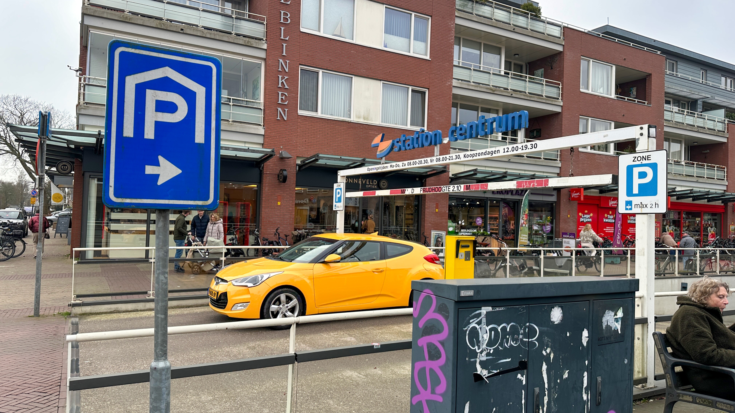 Geel voertuig geparkeerd voor een winkelcentrum met parkeersignalen; mensen lopen langs winkels op de achtergrond.
