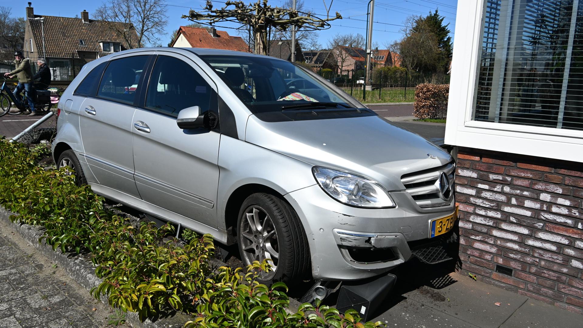 Een zilverkleurige auto is tegen een bakstenen muur en een tuinhek gebotst, met schade aan de bumper.