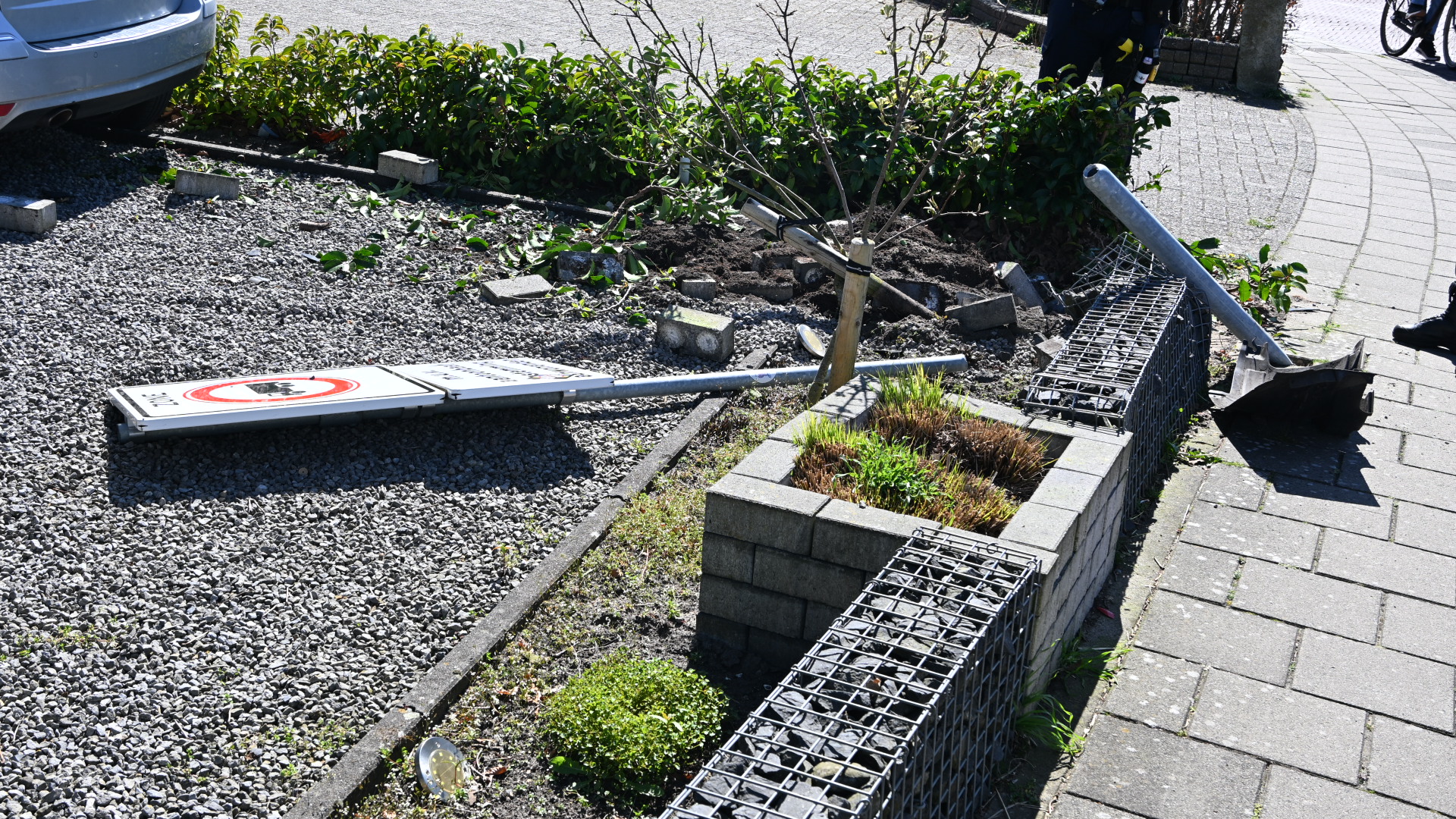 Omgevallen verkeersbord en beschadigde haag naast een geparkeerde auto en trottoir.
