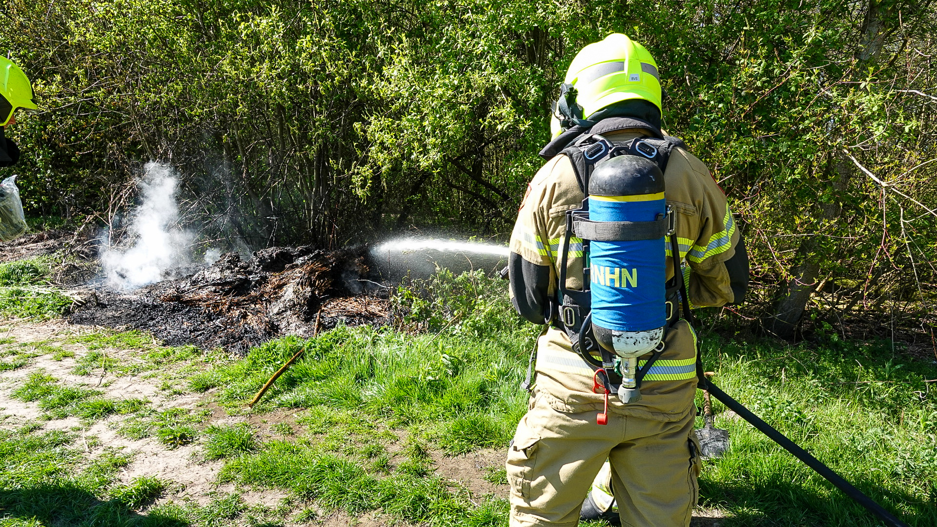 Brandweerman in beschermende kleding blust een smeulend stuk grond naast struiken.