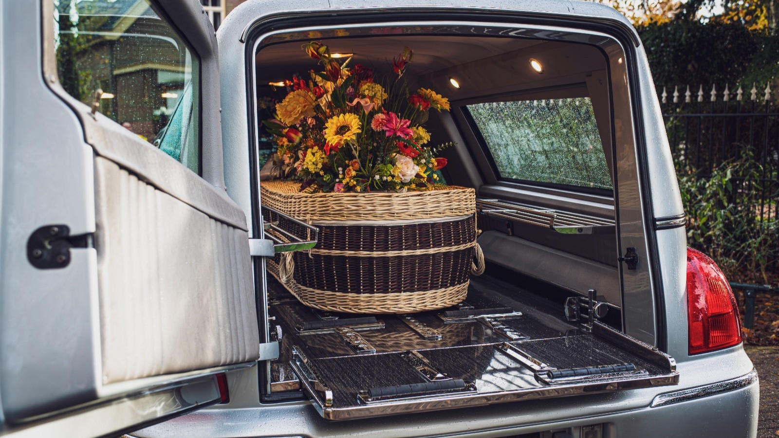 Gevlochten lijkkist met bloemen in de open achterkant van een zilveren begrafenisauto.